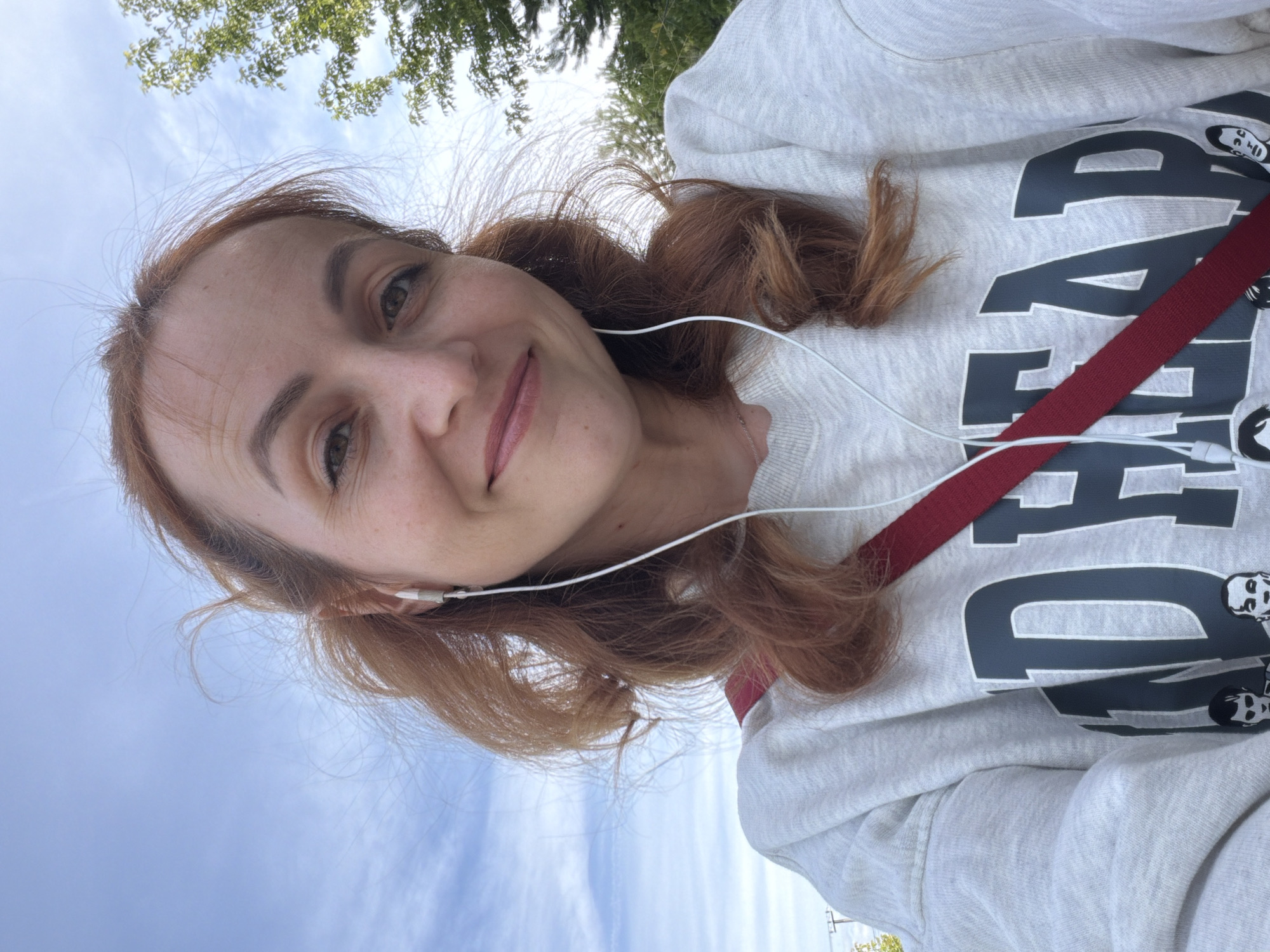 An outdoor, medium-shot selfie is taken from a low angle, looking up at a red-haired woman in her late 20s to early 30s. She has fair skin with freckles, light brown eyes, and is smiling slightly. Her hair is a medium reddish-brown, styled in loose waves, with some strands flying in the wind. She is wearing white earbuds and a light gray crewneck sweatshirt with a dark gray graphic on the front that appears to read "BAD..." with some faces below. A red strap is visible diagonally across her chest, suggesting she is wearing a bag. The background is a bright blue sky with thin, wispy clouds. To the right, a portion of a green tree is visible. The image appears to have been taken on a sunny day. The location in the city of Chaikovsky, Russia is not discernible from the provided image.
