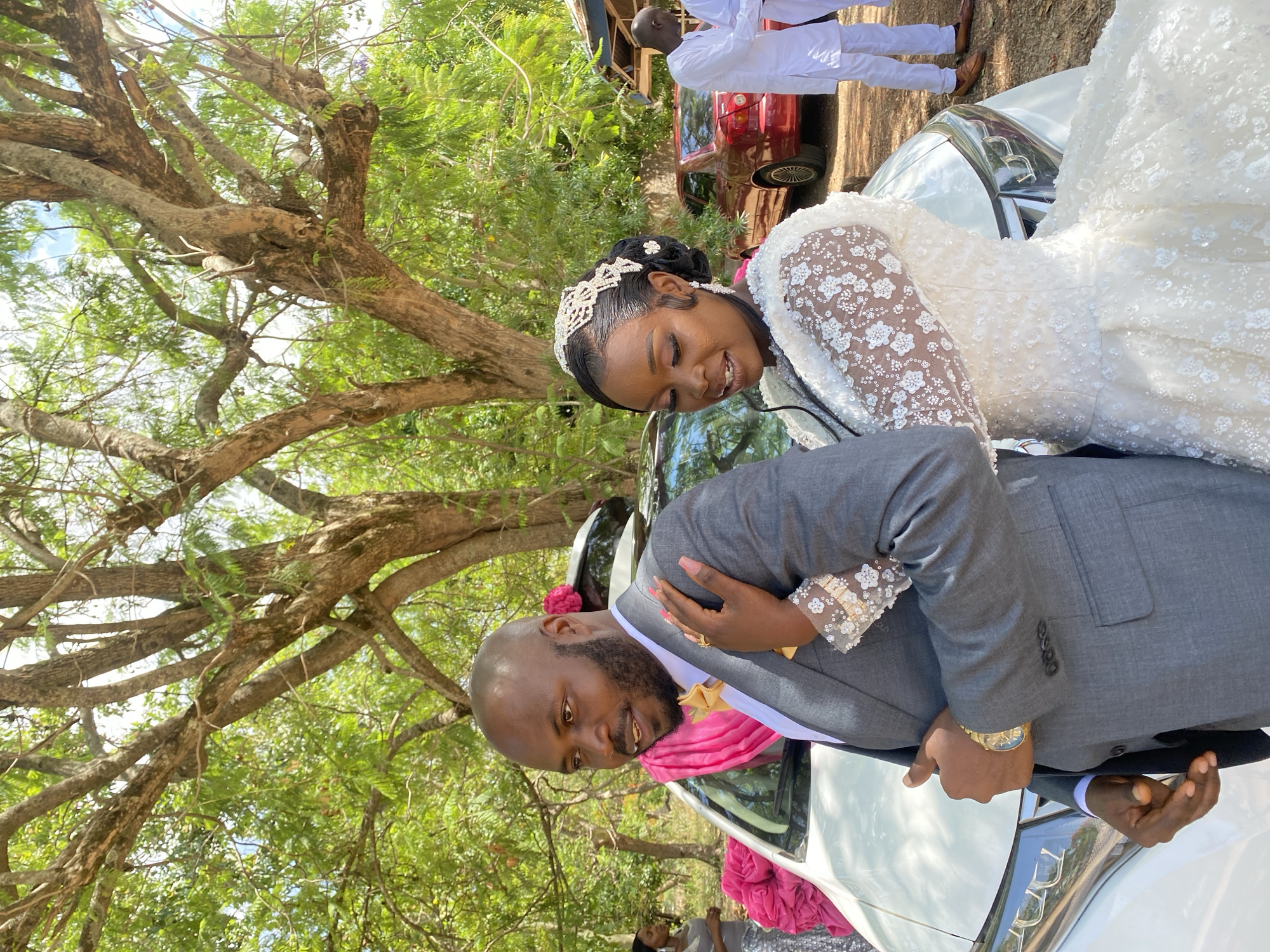 The image shows a wedding scene in Dutsen Kato, Nigeria. A groom in a grey suit and pink scarf, with a gold watch, is standing next to a bride in a white wedding gown with lace sleeves and a beaded headpiece. They are positioned next to a white car. Behind them, other individuals are visible, including one in a white outfit. A red car can also be seen. Trees with green leaves surround the scene.