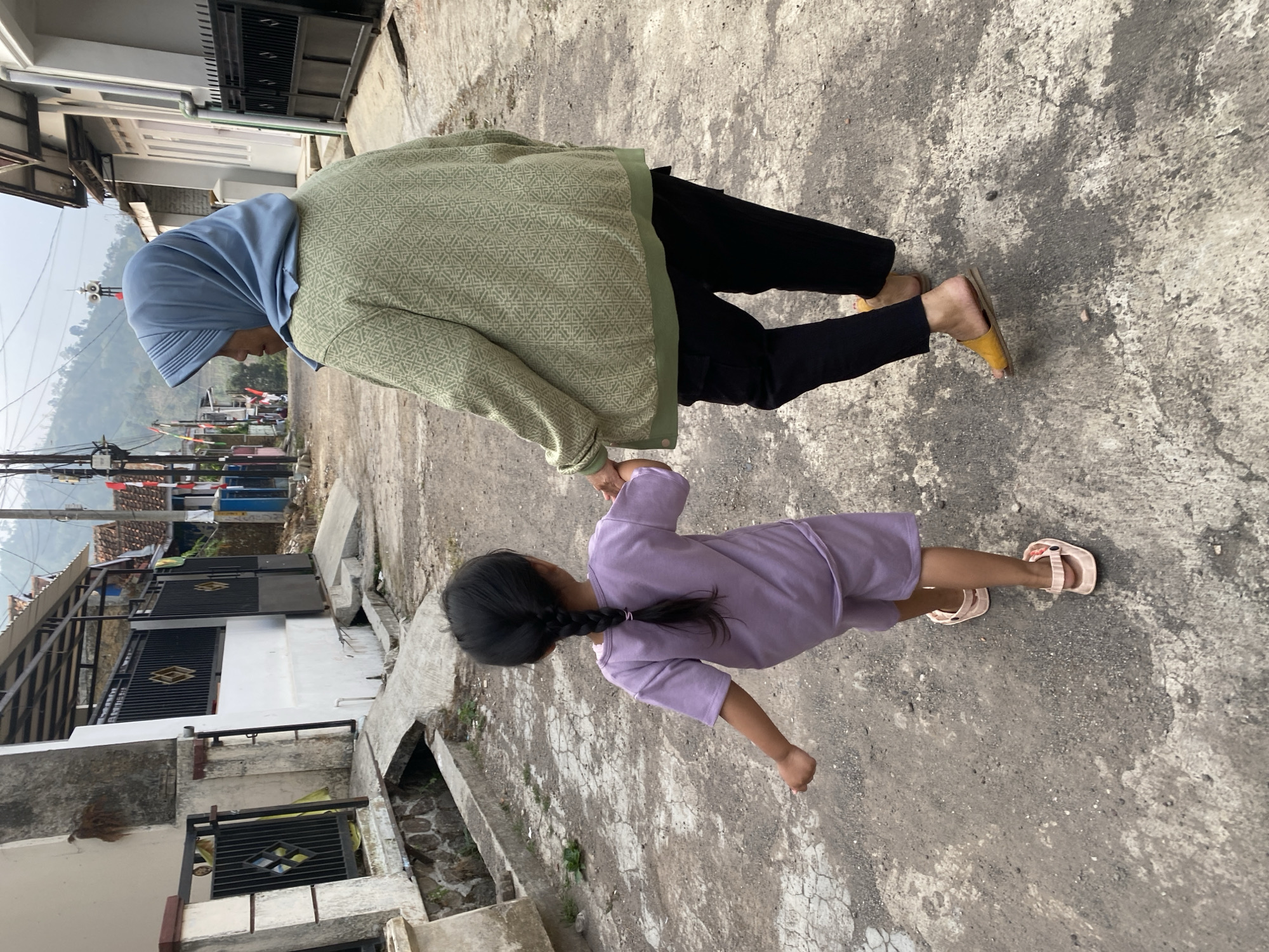 An Indonesian woman wearing a blue hijab and a green patterned sweater, and a young girl with her hair in a braid and wearing a purple tracksuit, walk hand-in-hand down a narrow, paved street lined with houses. The houses appear to be of modest construction, some with metal gates and awnings. In the background, a hillside is visible, suggesting a somewhat rural or semi-urban setting. The street itself is weathered and cracked, with some patches of dirt. The scene takes place in Garut, Indonesia, indicated by the presence of Indonesian flags along the street, which are common during national celebrations. The overall atmosphere is one of everyday life, with the woman and child enjoying a stroll.