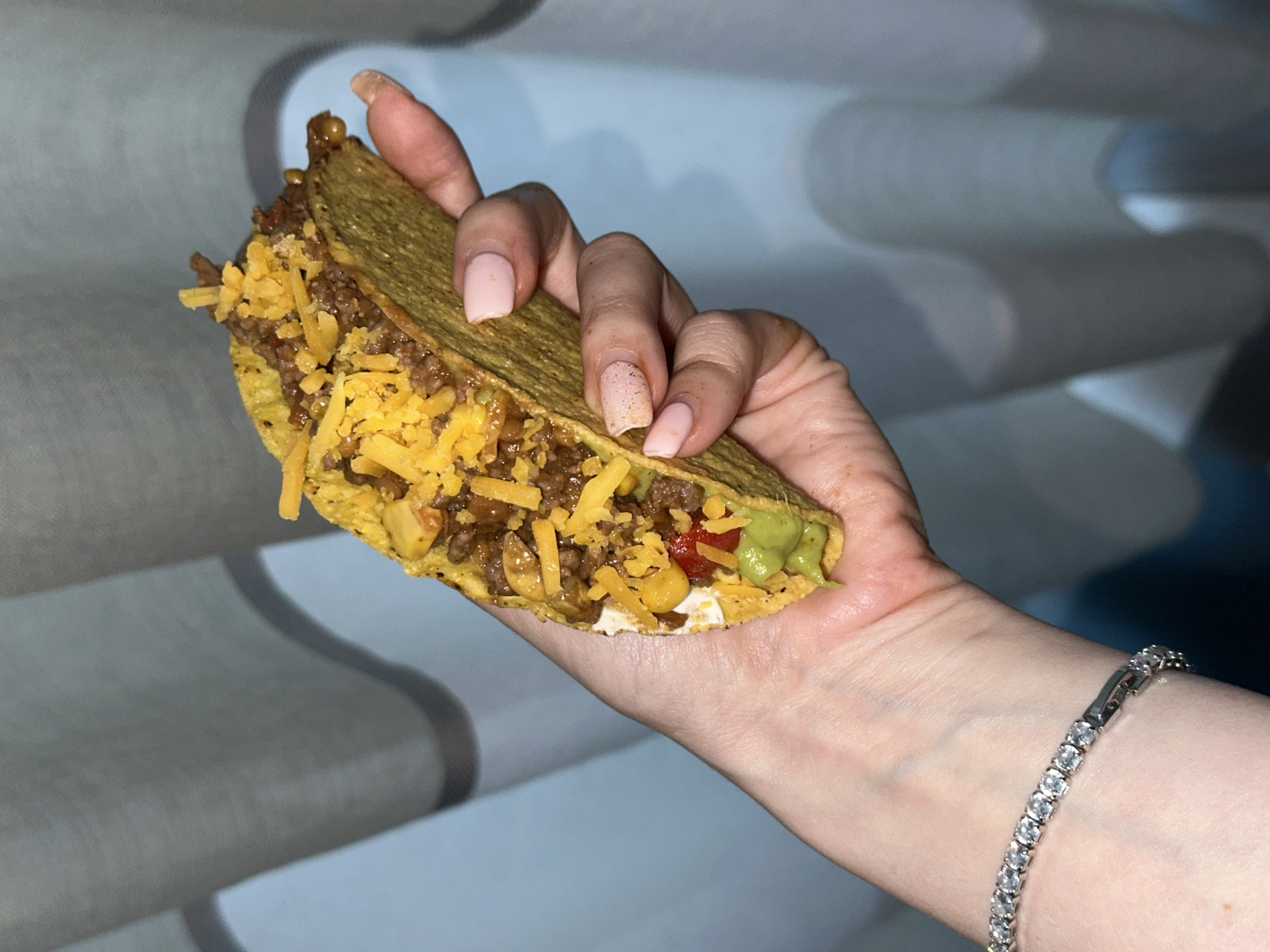A close-up shot captures a person's hand holding a hard-shell taco. The taco is generously filled with ground meat, shredded yellow cheese, some diced red ingredients (likely tomatoes), and a dollop of green guacamole peeking out from one end. The taco shell appears crispy and golden-brown.
The hand, which is fair-skinned, has fingernails painted with a light pink or nude polish. Some nails appear long, and one shows a slightly speckled or chipped finish. A sparkling, silver-toned bracelet featuring a continuous line of clear, possibly diamond or cubic zirconia, stones adorns the wrist.
The background consists of a light-colored, textured surface with distinct wavy or ridged patterns, resembling blinds, curtains, or a decorative wall panel. The lighting is somewhat dim, suggesting an indoor setting under artificial light. The activity depicted is simply the holding of the taco. No text is visible in the image. The location context for this image is Amsterdam, Netherlands.