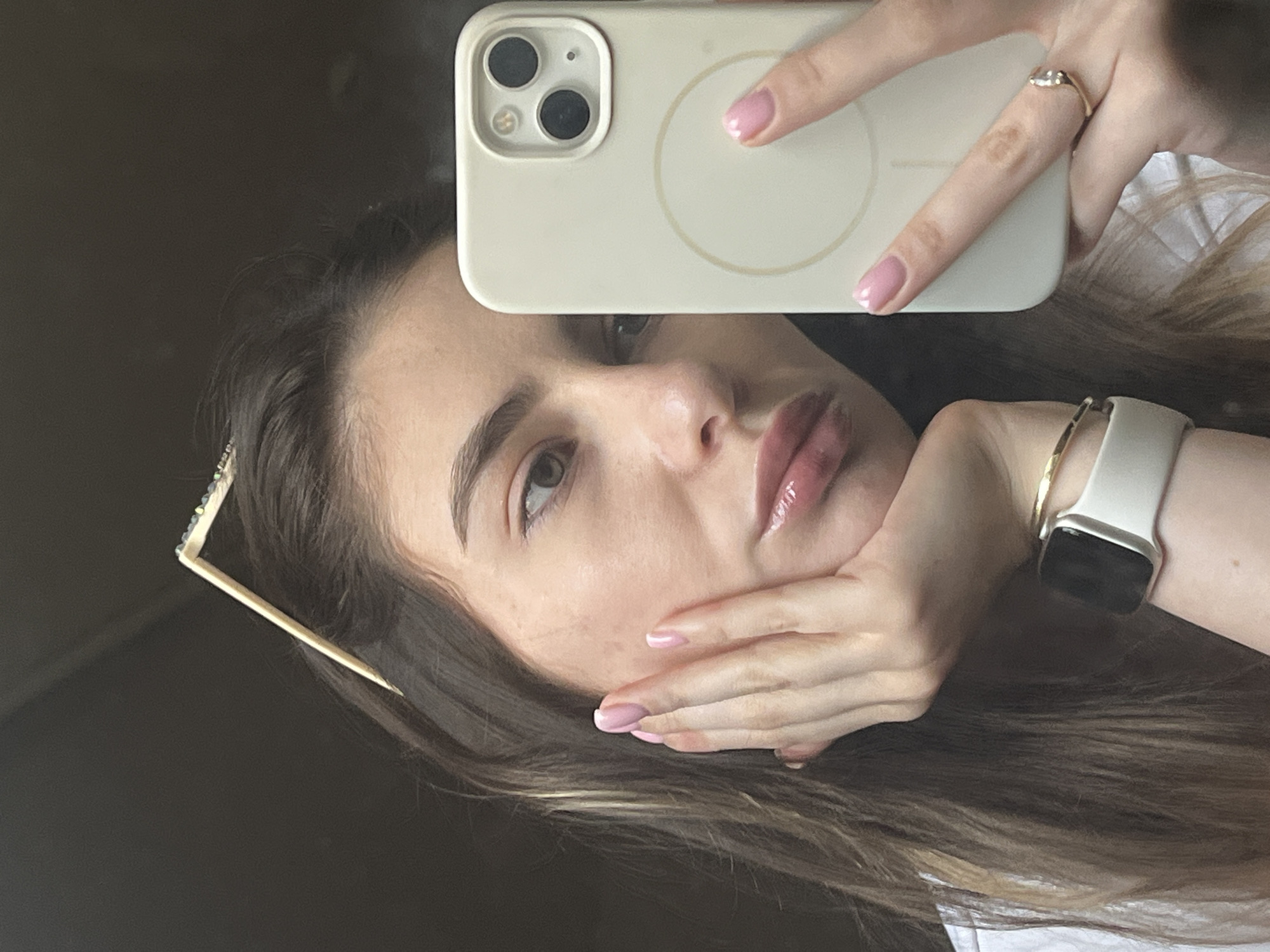 A young woman poses for a selfie. She holds a white iPhone with three camera lenses in her right hand, positioning it above her head. Her face is partially obscured by the phone. She has light skin, brown hair, and pink nail polish. She is wearing a gold ring and a white Apple Watch with a gold band. A barrette is visible in her hair. The background is dark. The location is Unknown, Unknown.