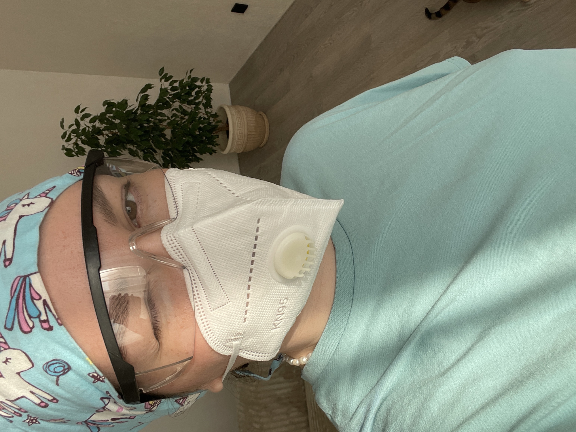 A close-up, chest-up selfie depicts an individual located in a residential setting in Izhevsk, Russia. The person is wearing a light blue scrub cap adorned with white unicorn and various cartoon patterns, clear safety glasses with black frames, and a white KN95 respirator mask featuring an exhalation valve. A pearl necklace is visible around the neck, and the person is dressed in a light blue short-sleeved t-shirt. The left eye is winked shut, displaying pronounced periorbital wrinkles, while the right eye is open and brown. The background includes a textured light-colored sofa or textile object to the left. To the right, a tall potted plant with dark green foliage is situated in a patterned beige pot. Light-colored walls converge in the background, above a light-toned wood laminate floor. A black electrical outlet is visible on the right wall, and a partial view of an orange and black striped object, consistent with a cat's tail, is visible on the floor at the bottom right.