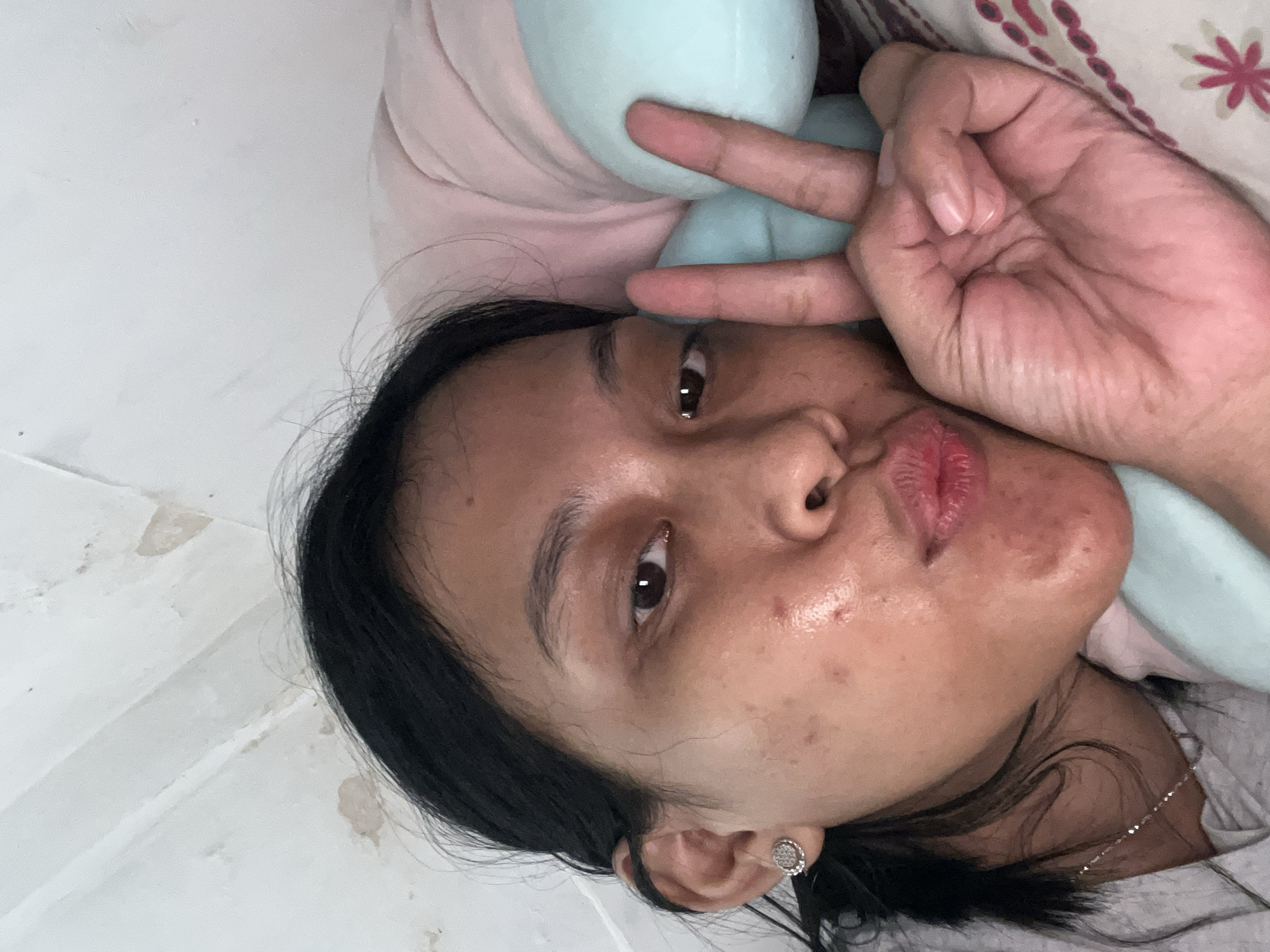 A young Indonesian woman with dark hair and brown eyes lies in bed, resting her head on a pillow and a stuffed animal. She is looking at the camera and puckering her lips, while making a peace sign with her right hand. She is wearing a grey t-shirt and a necklace. The background consists of a white ceiling with some water stains and a patterned bedsheet. The setting appears to be a bedroom in a home in Kecamatan Pati, Indonesia.