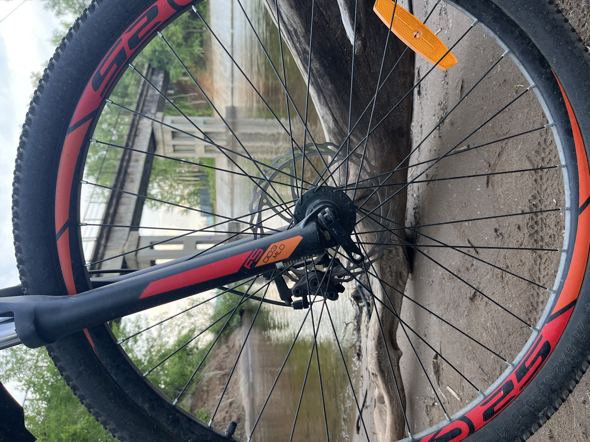 A close-up shot of a bicycle wheel. The wheel is black with a red and orange stripe. The wheel is resting on the ground, which appears to be sand. In the background, there is a body of water and a bridge. Based on the landscape and the specific bridge structure, the location can be identified as the city of Chaikovsky, Perm Krai, Russia. The bicycle wheel is likely leaning against a piece of driftwood. The focal point of the image is the bicycle wheel, with the background providing context to the location.