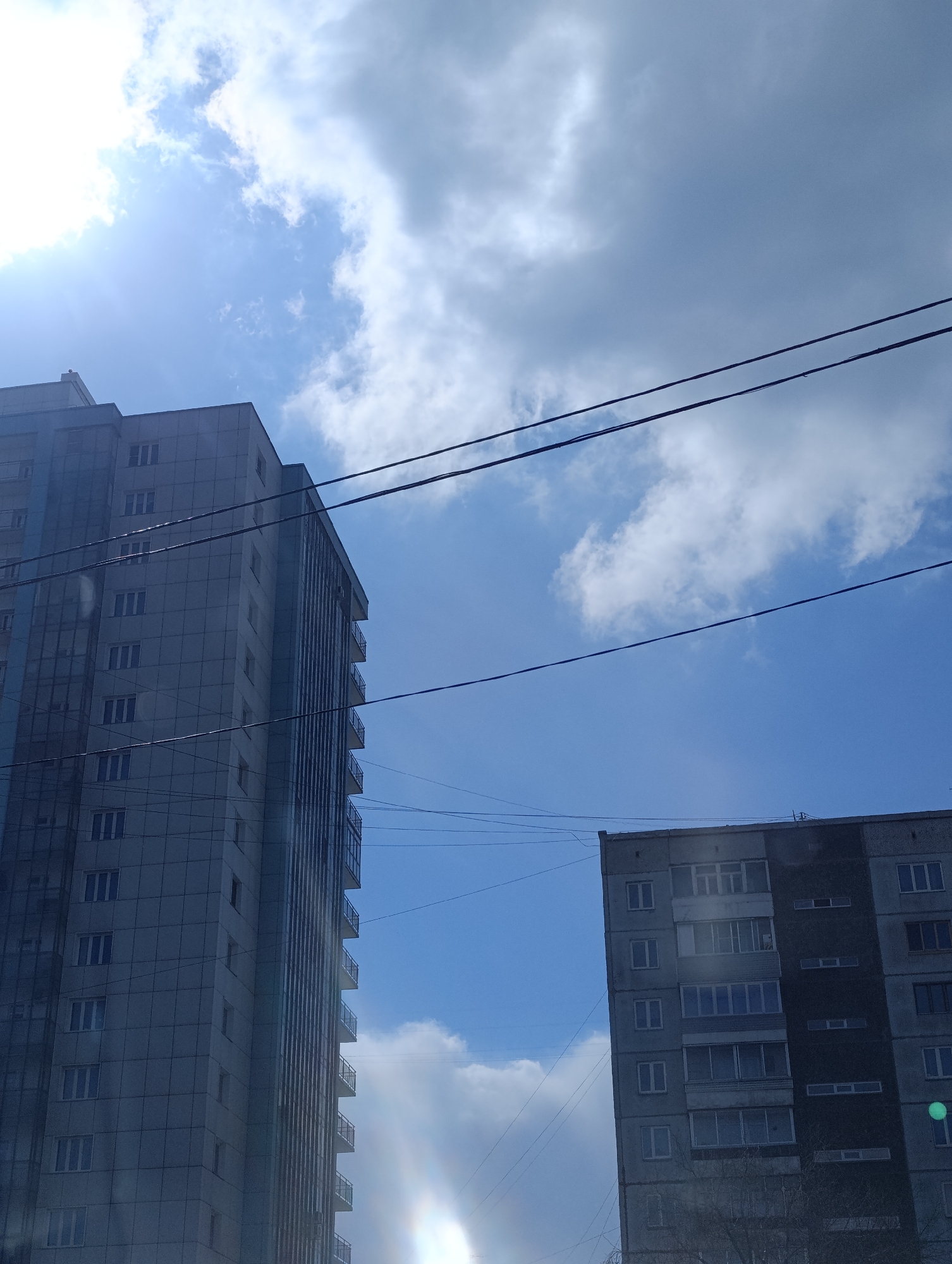 The image shows two buildings under a sky with scattered clouds in Krasnoyarsk, Russia. The building on the left is a modern high-rise with a metallic or glass exterior and numerous windows. The building on the right is an older, Soviet-era apartment block with a concrete facade. Power lines are visible stretching across the sky between the buildings. The sun is shining brightly, causing glare and some lens flare. There are no people visible.