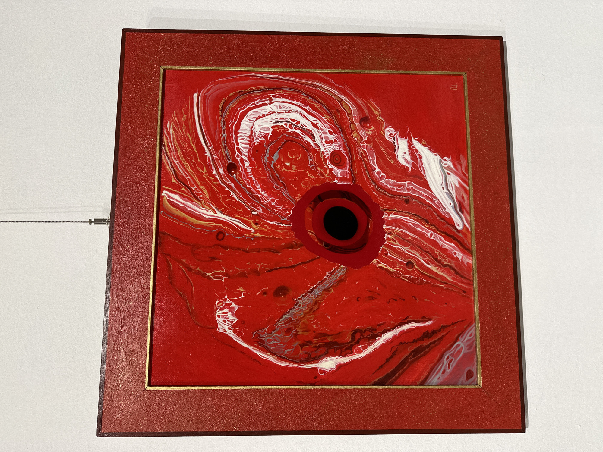 An abstract acrylic pour painting, predominantly red, is displayed centrally on a clean white wall, likely in an art gallery or home. The painting is framed in a textured red border with a thin golden inner trim.
The artwork features swirling and organic patterns created by fluid art techniques, with dominant hues of vibrant red, contrasting white, and shimmering gold. These colors flow and intermingle across the canvas, creating a dynamic, marbled effect with various cells and intricate lacing visible in the paint. At the core of the composition is a prominent black circle, surrounded by concentric rings of deep red, suggesting a focal point like an eye, a vortex, or an orifice. The fluid patterns emanate outwards from this central void, giving the impression of movement or an unfolding energy.
In the upper right corner of the painting, small, stylized initials or a signature are visible, possibly resembling "ALL." The white wall behind the painting is bare, with a thin, metallic art-hanging cable visible on the left side of the frame. The overall presentation is clean and focused on the artwork itself. The location context for this media file is Minsk, Belarus.