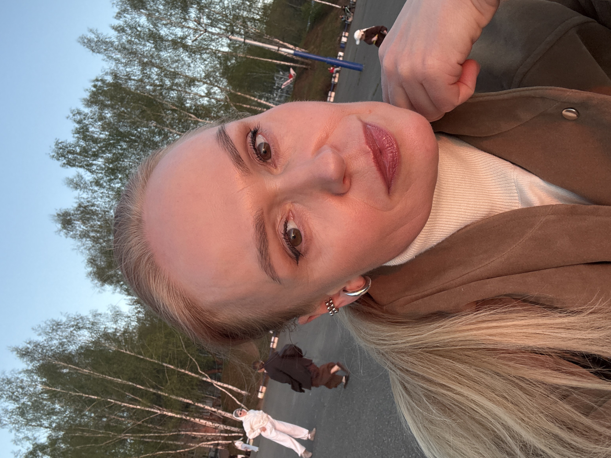 A selfie-style photograph shows a woman with blond hair, light skin, and light-colored eyes wearing a brown jacket and white turtleneck sweater. She has a silver hoop earring and another ornate earring in her left ear. Behind her, two people walk on a paved surface, one in a white outfit and the other in brown clothing. Trees with bare branches are visible in the background against a light blue sky. A blue and red structure is in the distance, possibly related to an athletic field. The scene may be in Chaikovsky, Russia.