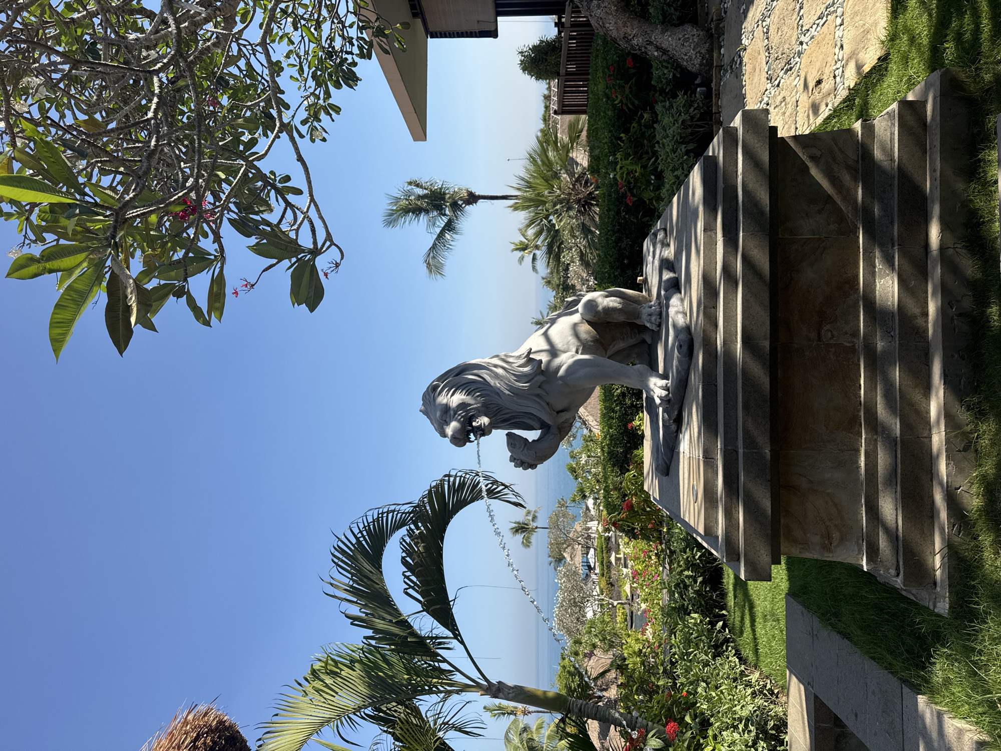 The photograph depicts an outdoor scene likely located in Kuta Selatan, Indonesia, given the presence of tropical vegetation and coastal views. A stone lion statue stands atop a series of stone steps, with the ocean visible in the background. The landscape includes palm trees, flowering bushes, and well-maintained grass. The sky is clear and blue, suggesting a sunny day. The statue appears to be a decorative feature within a garden or landscaped area of a hotel. The presence of a building structure partially visible on the left side of the frame suggests an architectural setting.