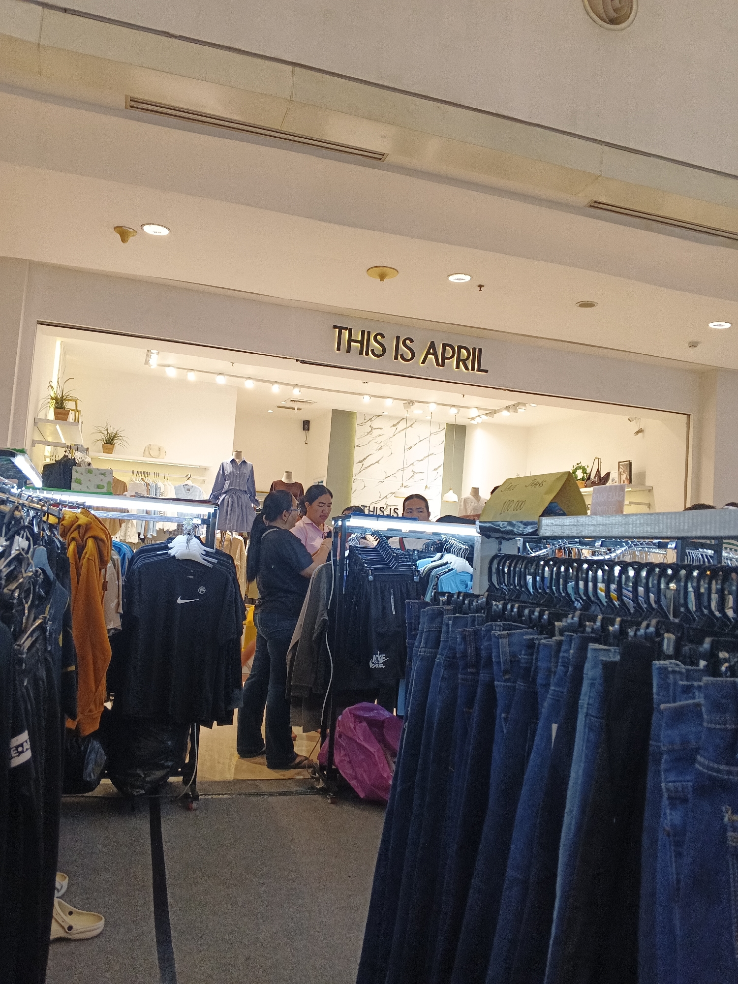 In City of Medan, Indonesia, a clothing store named "THIS IS APRIL" is depicted. The store features a well-lit interior with displays of apparel. In the foreground, a rack of dark blue denim jeans is prominently displayed. Further inside, other clothing items, including shirts and jackets, are visible on hangers. Two women are present inside the store; one, wearing a pink top and dark pants, appears to be interacting with the other woman who is wearing a black shirt and glasses. In the background, mannequins are dressed in fashion items. The store's branding, "THIS IS APRIL," is visible above the entrance. The flooring inside the store appears to be wooden, while the aisle area is carpeted with a dark grey material. A bright pink duffel bag is situated on the floor between the clothing racks.
