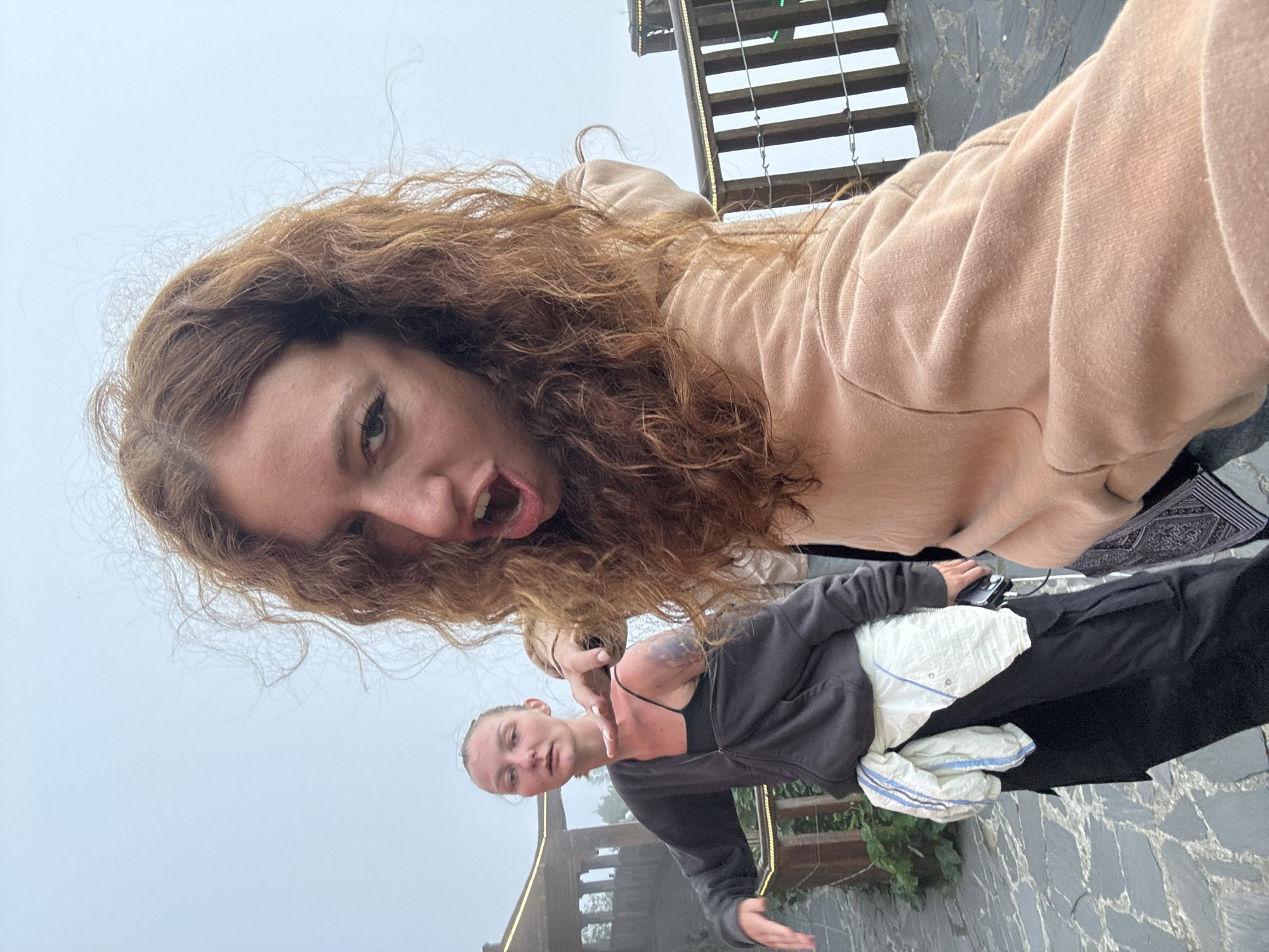 The image shows two Caucasian women taking a selfie outdoors, likely in Binh Lu, Vietnam, based on the mountainous, cloudy landscape visible in the background. One woman is positioned closer to the camera, with curly, reddish-brown hair, wearing a light pink hoodie, and making a funny face with her mouth open. The second woman is standing behind her, wearing a dark gray zip-up hoodie over a black tank top, dark pants, and holding a white and blue striped towel. The ground is paved with gray stone tiles, and a railing is visible behind the women. The sky is overcast.