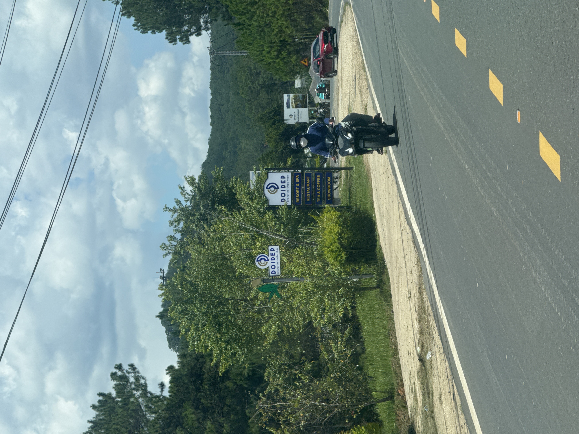 The image shows a road in Thôn 6, Vietnam, with a motorcyclist riding on the right side of the frame. The road is paved with asphalt and has yellow lane markings. On the left side of the road, there is a sign for "DOIDEP" which appears to be a resort and spa, restaurant, tea & coffee shop, and showroom. There are also trees and foliage on either side of the road. In the background, there are hills covered with trees. The sky is cloudy. The motorcyclist is wearing a helmet and a mask, and is riding a black motorcycle. There are other vehicles in the distance, including a red car and a few other motorcycles. The scene appears to be captured during the daytime, with moderate lighting.