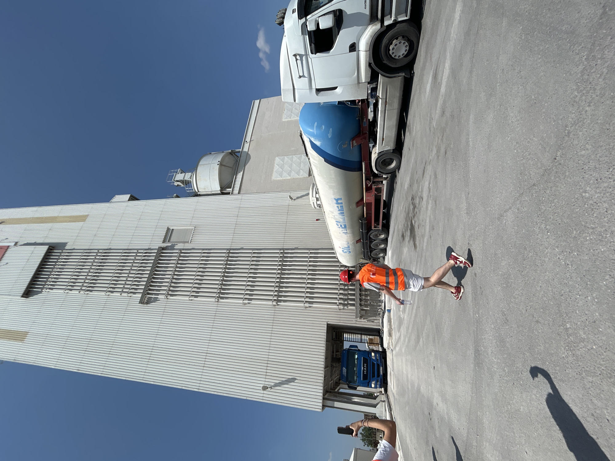 The image captures an outdoor industrial scene under a clear blue sky, likely in Elin Pelin, Bulgaria. Dominating the left side of the frame is a tall, modern, corrugated metal building with vertical elements. In the mid-ground, a blue MAN truck is partially visible entering an opening in the building. To the right, a white semi-trailer truck with a large, blue cylindrical tank is parked. The words "SILO" and "ELMER" are visible on the side of the tank. A person wearing a red hard hat and an orange safety vest walks across the paved area, carrying a bottle of water. Another person is partially visible on the far left, holding up a smartphone, possibly taking a photograph. The ground is a rough, grey asphalt surface. A prominent shadow of a person with an outstretched arm is cast on the ground to the lower left. The scene appears to be a logistics or industrial processing facility.