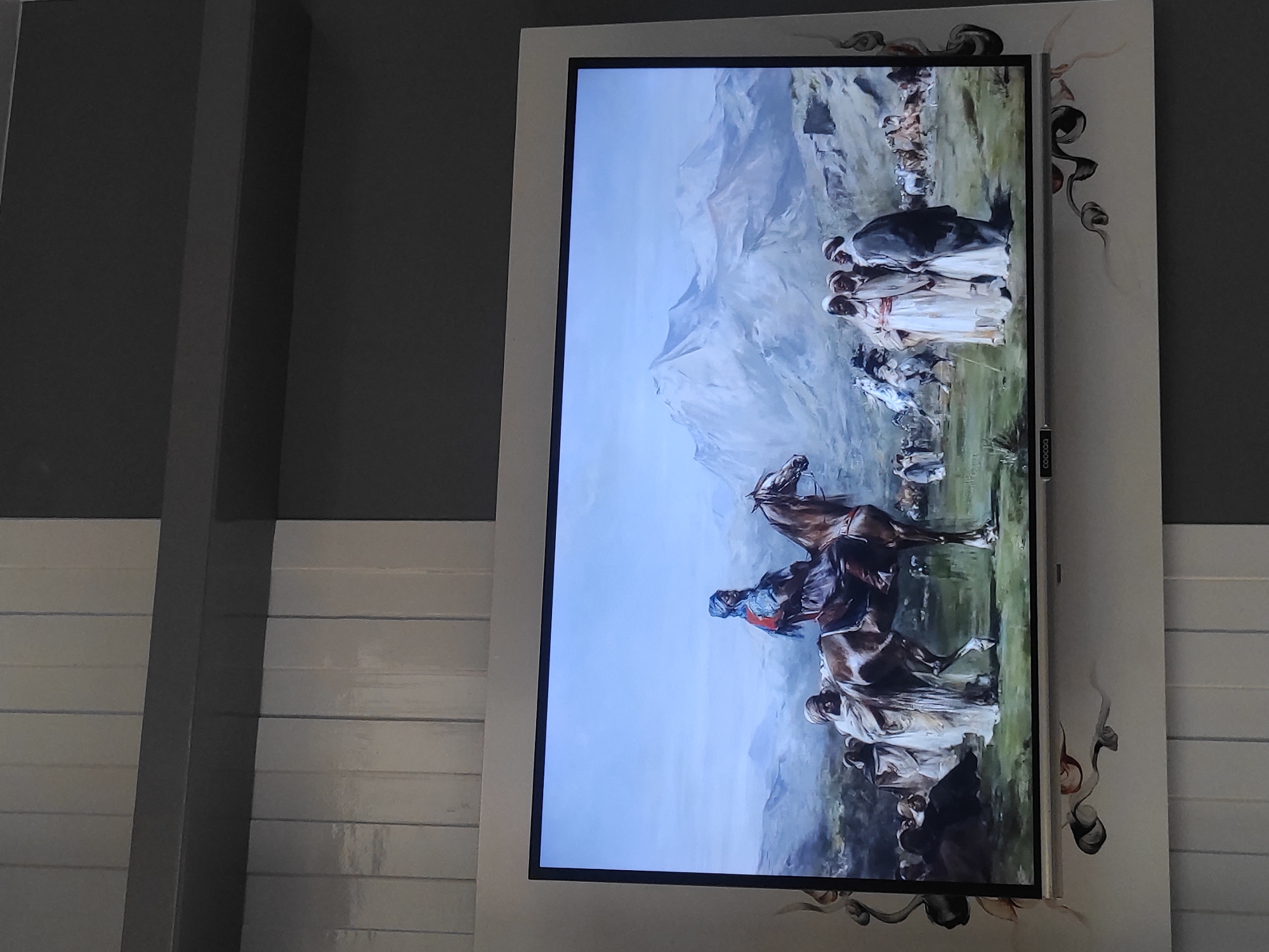 The image shows a large television screen displaying a painting of a historical scene. The painting depicts a man on horseback wearing a blue turban and a red garment. He is surrounded by a group of people in traditional Middle Eastern clothing, some of whom are also on horseback. In the background, there are mountains and a vast, open landscape. The television is mounted on a wall decorated with abstract swirling patterns.
The specific location of this television screen in the city of Sumedang, Indonesia, is not determinable from the provided image.
