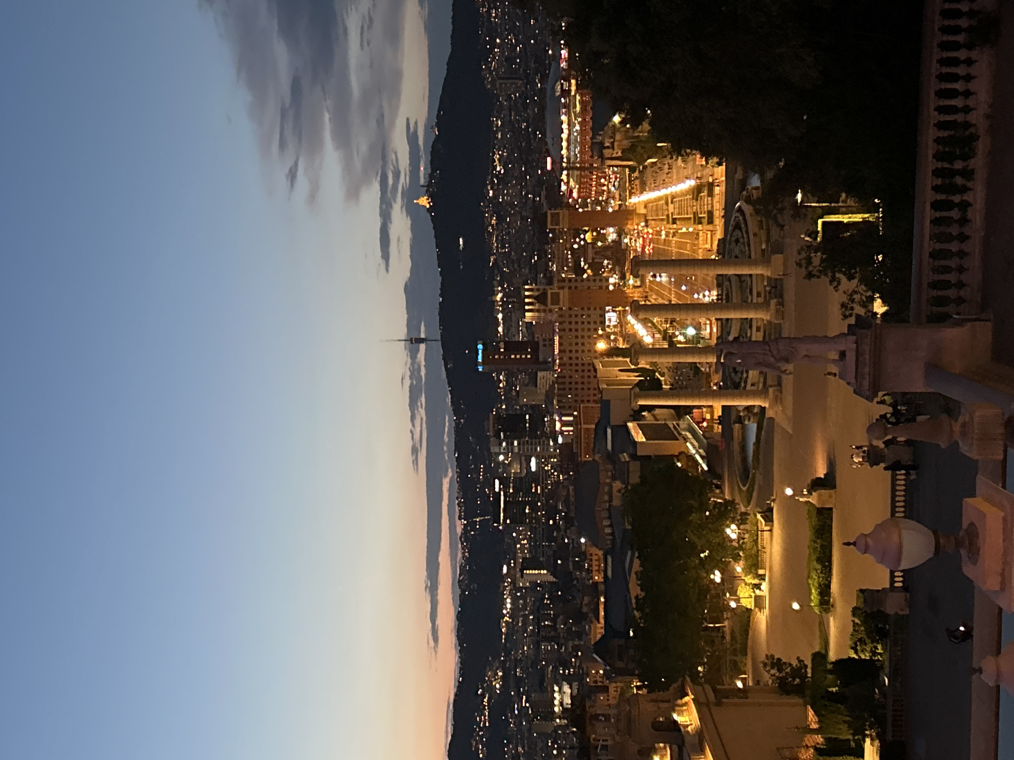 A panoramic cityscape of Barcelona, Spain, unfolds at dusk or early evening, seen from an elevated vantage point, likely Montjuïc, looking towards Tibidabo. The sky transitions from a soft blue at the top left to a faint yellowish hue near the horizon, with darker grey clouds clustered on the right.
The distant landscape features a range of mountains, along which the city lights extend. Two prominent structures punctuate the distant skyline: a tall, slender telecommunications tower (Torre de Collserola) is visible on the left side of the distant cityscape, with an "Allianz" logo subtly visible on a building directly beneath it. Further to the right, on a hilltop, the distinctly illuminated Temple Expiatori del Sagrat Cor church stands out.
The city below is a tapestry of countless illuminated buildings, with streets and structures glowing with artificial light. In the immediate foreground on the right, grand architectural elements are visible, likely part of the Palau Nacional or similar monumental structures. These include large classical columns, portions of staircases, and statues. Ornate street lamps and other outdoor light fixtures cast warm glows, illuminating walkways and small patches of trees and dark foliage. No specific human activity is discernible, and no individuals are clearly visible. The overall scene captures the expansive and serene beauty of Barcelona transitioning from twilight to night.