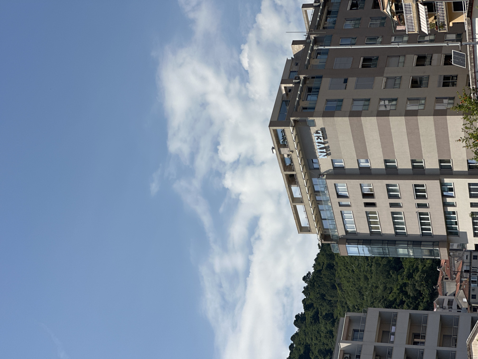 The image shows a view of a building in Budva, Montenegro, identified by the word "TRECAN" vertically displayed on its facade. The building has a striped design, with alternating light and dark tan vertical sections. It is a multi-story building with multiple windows. A forest is visible behind the building. Part of another building is seen in the lower right corner of the image. The sky is light blue with white clouds, taking up most of the image. The orientation of the image is sideways.