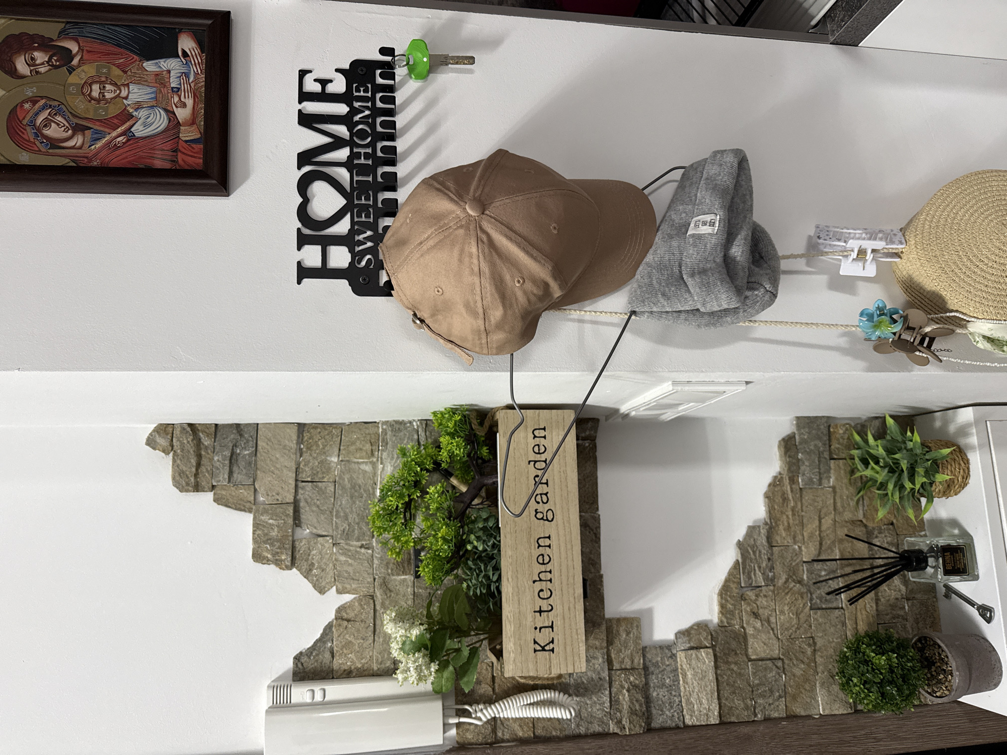 A wall in what appears to be a home in Sofia, Bulgaria is shown. The wall is white, with sections of decorative stone affixed to it. A framed religious icon is on the top left, and next to it a black metal wall-mounted key holder shaped like the word "HOME SWEET HOME," with a green-handled key hanging from it. Three hats are hanging on hooks to the right of the key holder: a tan baseball cap, a gray beanie, and a straw hat. Below the hats, a wooden sign reading "Kitchen garden" hangs, next to a small potted plant. To the right, another potted plant and an air freshener are visible. A white intercom system is attached to the wall on the lower left. The scene is brightly lit, indicating an interior setting.