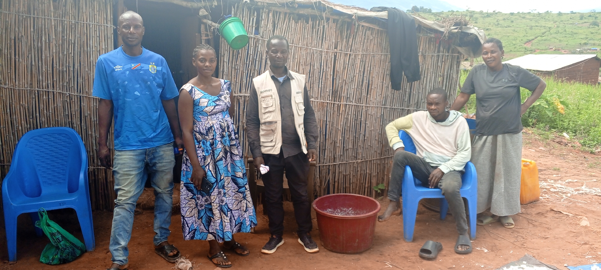 Outside a rudimentary dwelling with walls of woven sticks, five individuals stand or sit. A man in a blue soccer jersey and jeans stands to the left, next to a blue plastic chair with a green bag beneath it. A woman in a blue patterned dress stands beside him, holding a phone. To her right, a man in a vest stands, next to a large red plastic tub filled with small items. A man sits in a blue plastic chair, with a sandal on the ground near him. A woman in a gray shirt and long skirt stands with her hand on the chair. The environment appears to be a rural area, with a hillside and simple buildings visible in the background. This image was taken in the city of Makungu, Democratic Republic of the Congo.