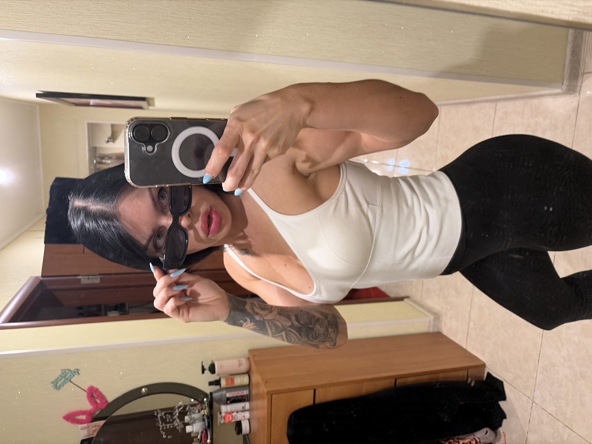 A woman is taking a selfie in a mirror in a room. She has dark hair, is wearing sunglasses, a white tank top, and black leggings. She has tattoos on her arm and is holding a phone in her right hand. In the background, there is a dresser with items on it, a doorway, and a framed picture on the wall. The room appears to be a bedroom or dressing area, with tiled flooring. There is no specific information in the image to indicate the location is in the city of Ufa, Russia.