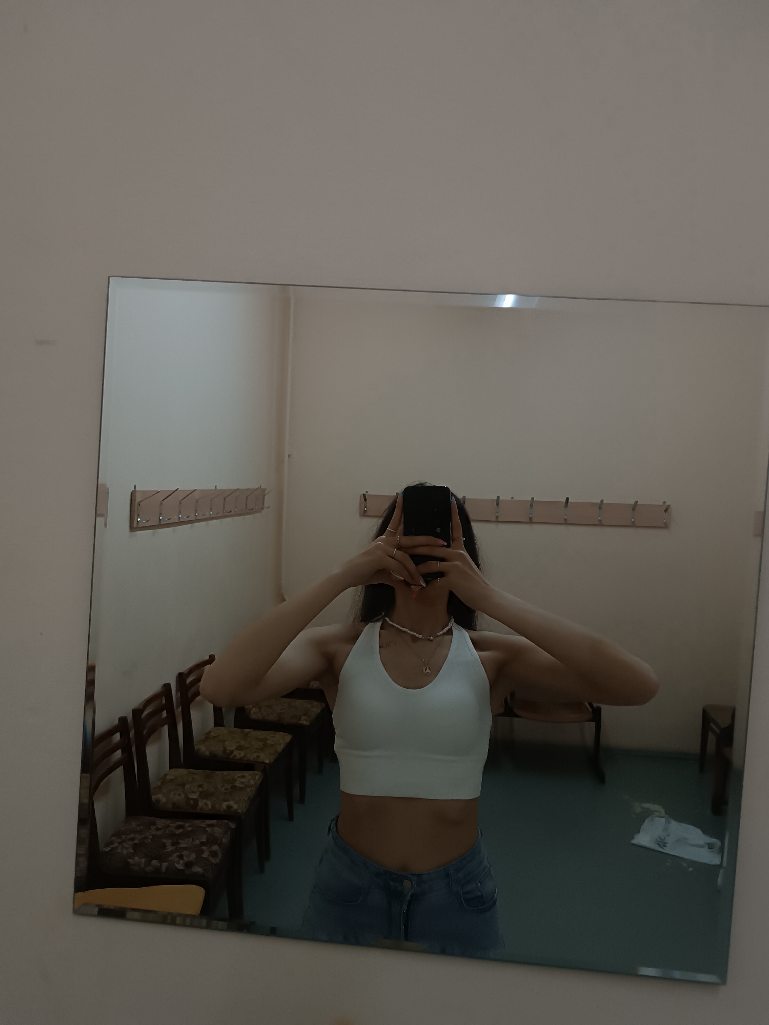 The image shows a person reflected in a mirror, holding a smartphone up to their face, obscuring it. The person is wearing a white halter top and blue jeans. The setting appears to be a dressing room, with wooden chairs and a row of coat hooks visible on the wall. The floor is a dull teal color. Location: Unknown, Unknown.