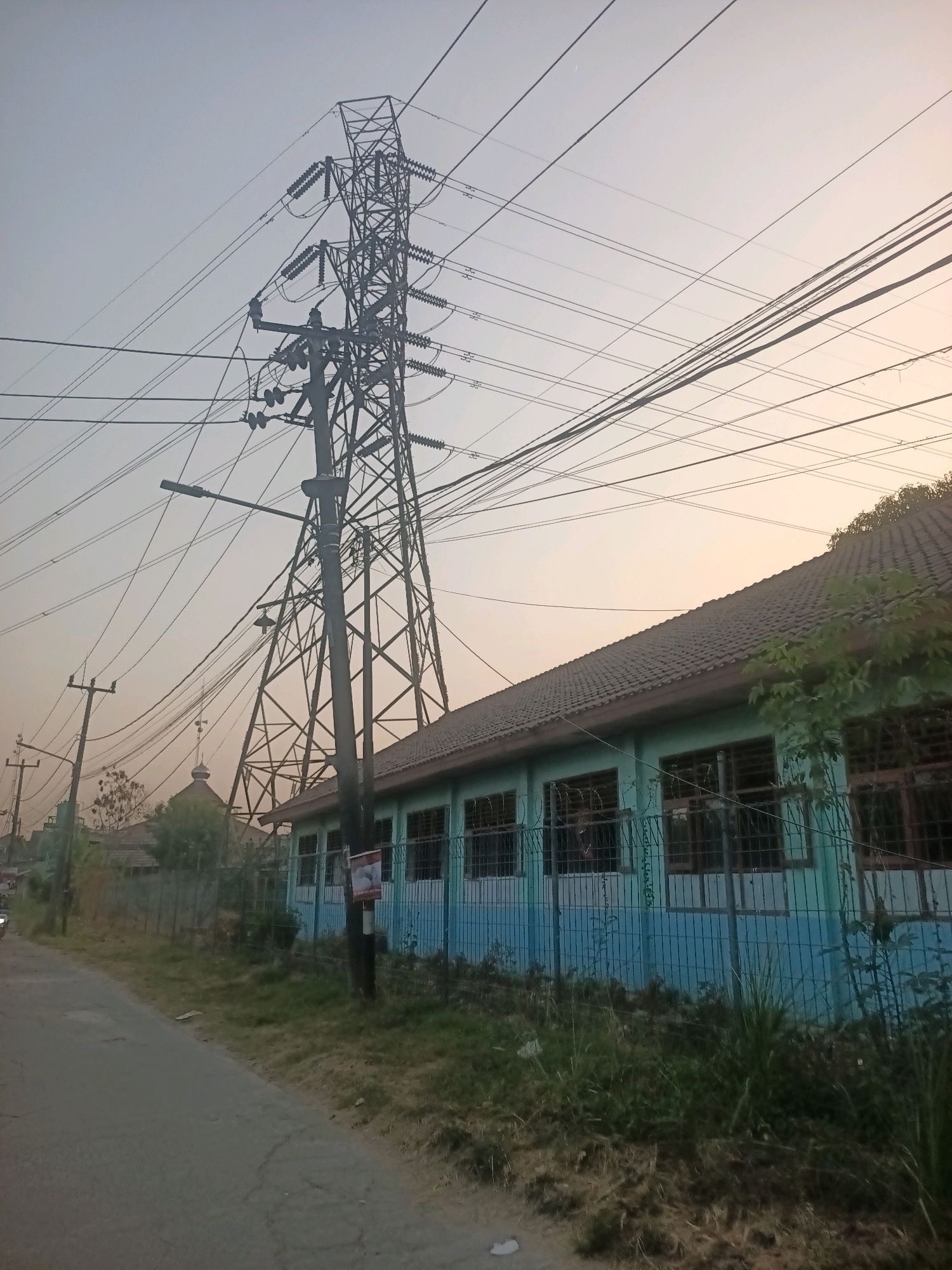 The image depicts an outdoor scene on a street in Wanajaya, Indonesia, during daylight hours, possibly late afternoon due to the sky's color. Dominating the center and right of the frame is a large, lattice-style electrical transmission tower, supporting numerous power lines that stretch across the sky. To the right of the tower, a long, low-rise building with a tiled roof and light blue and white walls is visible. This building has a series of windows, many of which are barred. A fence runs along the front of the building, partially obscured by vegetation.
To the left of the transmission tower, a paved road runs diagonally away from the viewer, bordered by a grassy verge. Several utility poles with electrical wires are also present along the road. In the distance, behind the building and a smaller utility pole, the dome of a building, possibly a mosque, can be seen, along with other structures and trees. The sky is mostly clear with a gradient of pale blue and warm, light orange hues, suggesting the sun is low on the horizon. There are no visible people in the image. The overall atmosphere suggests a semi-urban or suburban environment with significant electrical infrastructure.