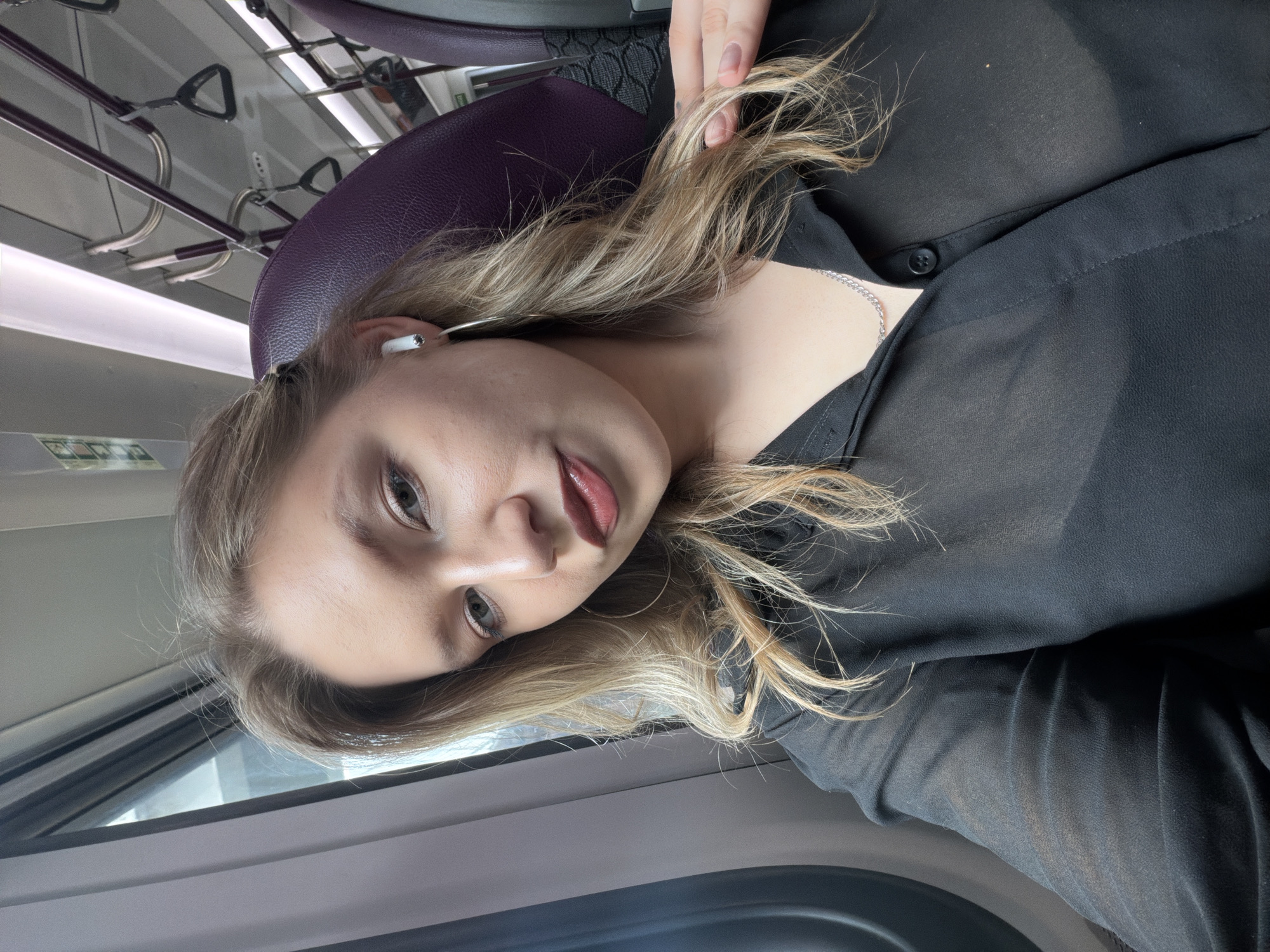 A blonde woman with medium-length wavy hair and fair skin is taking a selfie in a train car. She is wearing a sheer black button-down shirt, a silver chain necklace, and large silver hoop earrings. She has on white wireless earbuds and her makeup includes dark lipstick and eye makeup. The train car has purple upholstered seats and overhead handrails. The environment appears to be a public transportation setting, likely a train, with a window on the left side of the frame showing an overcast sky. The image captures the woman from the chest up, with a slight tilt of her head and a hand resting on her hair.