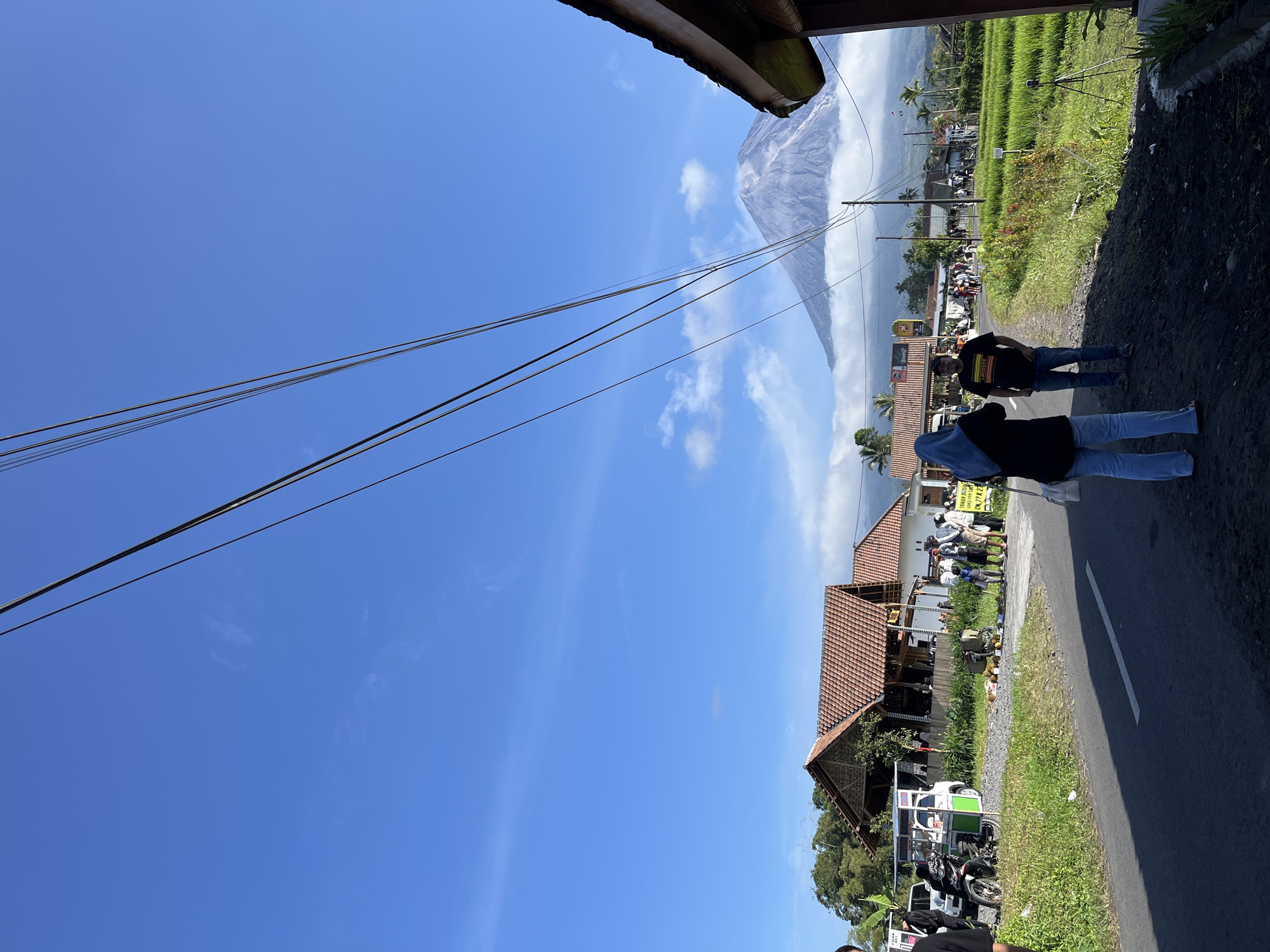 In a rural scene in Oro-oro Ombo, Indonesia, a conical volcano, Mount Merapi, looms in the background, its peak shrouded in clouds, with a plume of ash rising from its summit. The foreground features a paved road and a grassy verge. Several people are present, some walking along the road, others gathered near a food stall or small shop with a thatched roof. A motorcycle is parked on the side of the road. The sky is blue with scattered white clouds, and utility wires stretch across the frame. The environment appears to be a village with houses and rice paddies.