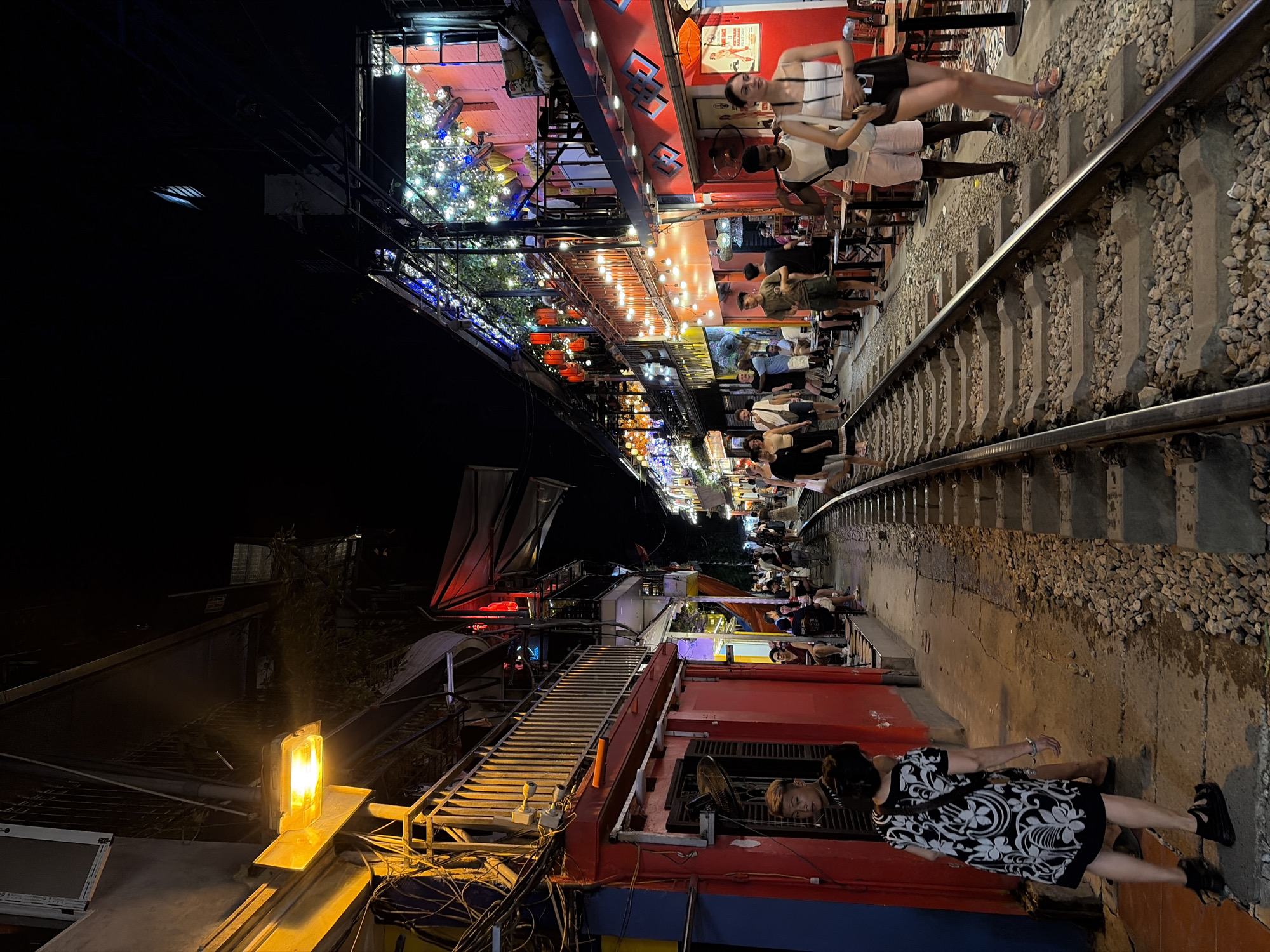The image depicts a narrow street in Hanoi, Vietnam, at night, with active train tracks running through the center. On either side of the tracks are brightly lit buildings, many of which appear to be cafes or restaurants, adorned with string lights and lanterns. People are seen walking along the sidewalk next to the tracks and sitting at outdoor tables. The architecture suggests a densely populated urban environment. The scene is lively, with a mix of locals and tourists enjoying the evening.