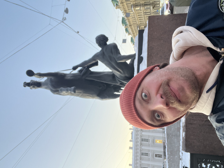 A man with a mustache and an orange knit beanie is depicted in a close-up selfie, looking directly at the camera with wide, slightly crossed eyes and a faint smile. He wears a dark jacket over a light-colored hooded top. Behind him, partially obscured by his head, is a bronze statue depicting a rearing horse with a struggling, nude male figure attempting to control it. This statue is one of the four "Horse Tamers" by Baron Peter Klodt, located on the Anichkov Bridge over the Fontanka River in Saint Petersburg, Russia.
Snow lightly dusts the dark red granite base of the statue. In the background, under a pale blue sky, a classical-style building with columns is visible to the left, and other historic multi-story buildings with pitched roofs, some green, line what appears to be a street or embankment to the right. Overhead electrical wires crisscross the sky. A vehicle is partially visible on the far right street. The scene is illuminated by natural daylight, indicating clear weather.