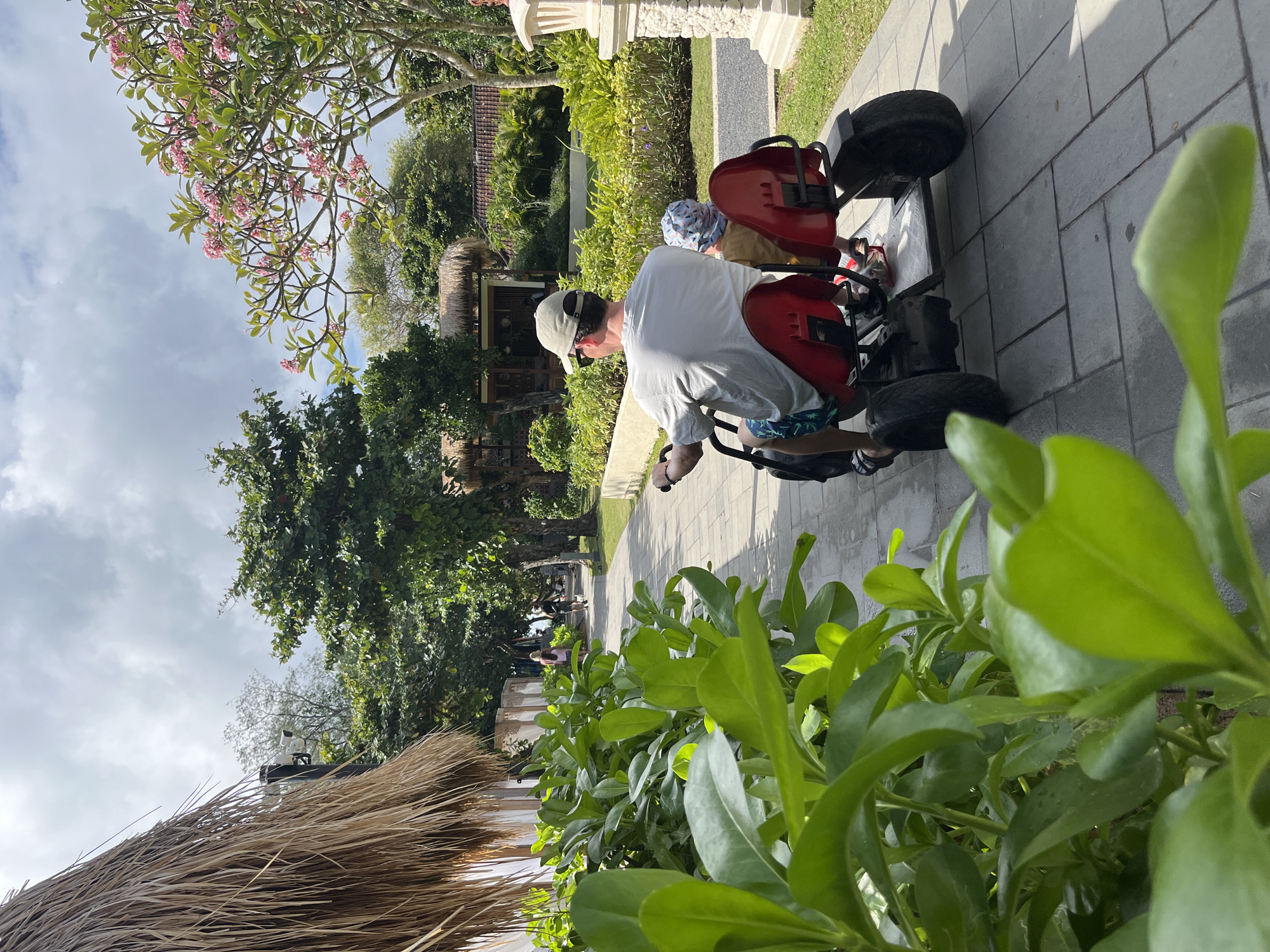An overhead shot captures a man and a child on a red electric cart on a paved pathway in Kuta Selatan, Indonesia. The man, wearing a gray t-shirt and a beige cap, drives the cart, while the child, in a patterned cap, sits behind him. Lush green foliage frames the foreground, and a thatched-roof structure is visible to the left. In the background, trees and other thatched-roof buildings suggest a tropical resort or village setting. The pathway is bordered by manicured grass and a light-colored wall. The sky is partly cloudy.