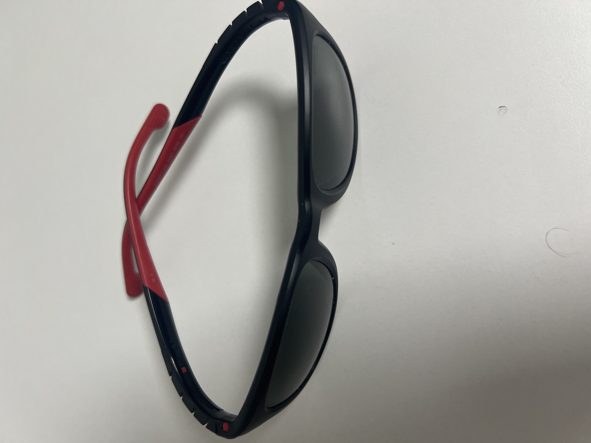 An overhead shot shows a pair of black and red sunglasses lying on a white surface. The sunglasses have dark lenses and black frames with red accents on the temples and bridge. The temples are partially extended, revealing the red inner lining and some inscribed text. The background is a plain white surface.