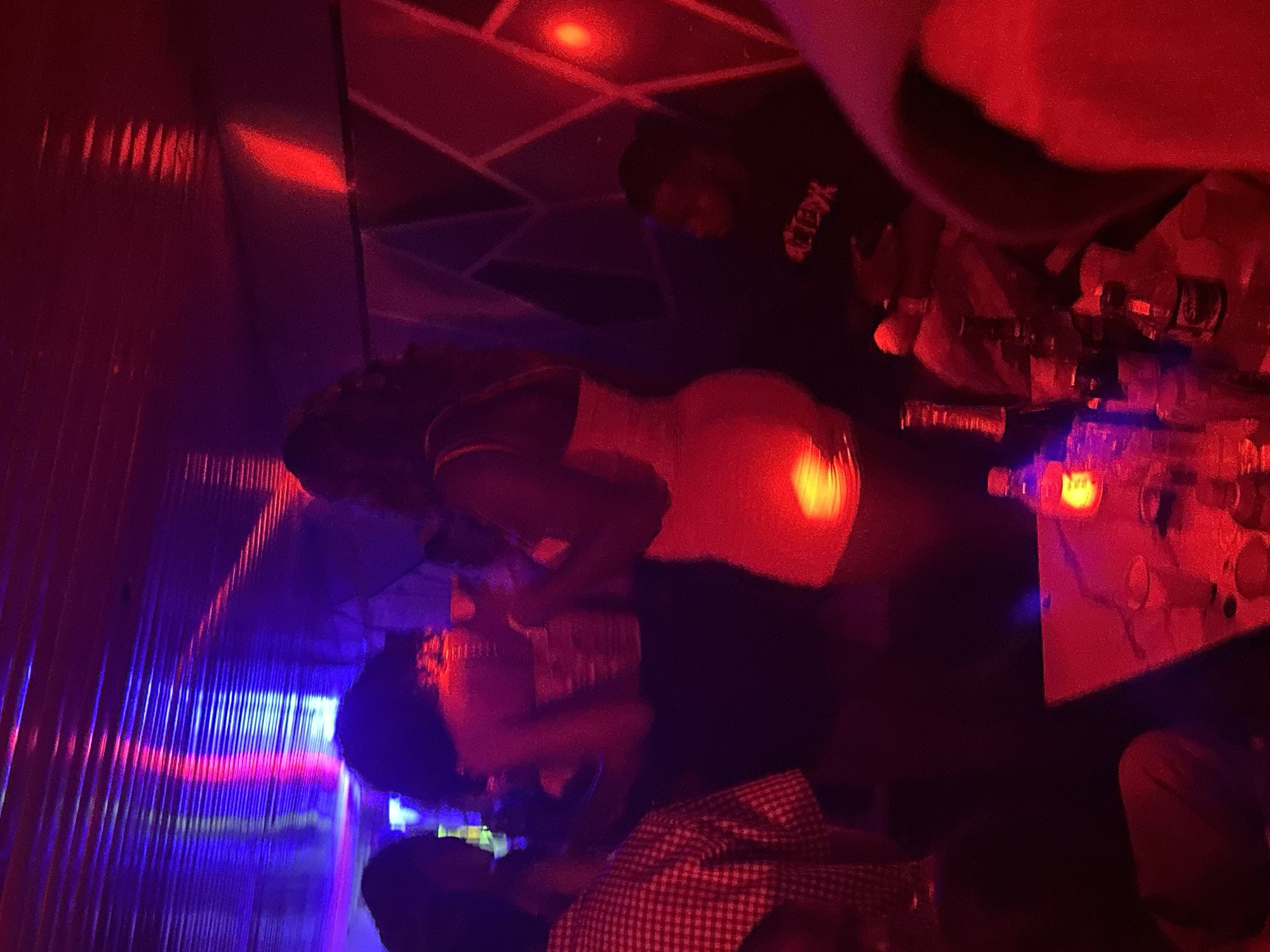 The image depicts a dimly lit interior, likely a nightclub or bar, bathed in red and blue stage lighting, with a metallic ceiling. In the foreground, a table is visible with several bottles of drinks, including one with a yellow label and another with a prominent white label and red writing. A person is seated in the lower right, partially visible, wearing a black hooded sweatshirt with white lettering. To the left, a person in a red and white checkered shirt is also present. In the mid-ground, two women are dancing. One woman, on the left, is wearing a strapless top and dark pants, while the other, in the center, is wearing a light-colored, form-fitting dress. Their poses suggest they are actively engaged in dancing. The overall atmosphere is that of a vibrant social gathering. The scene is consistent with nightlife establishments found in cities like Wumat, Nigeria, where such venues are common for entertainment and social interaction.