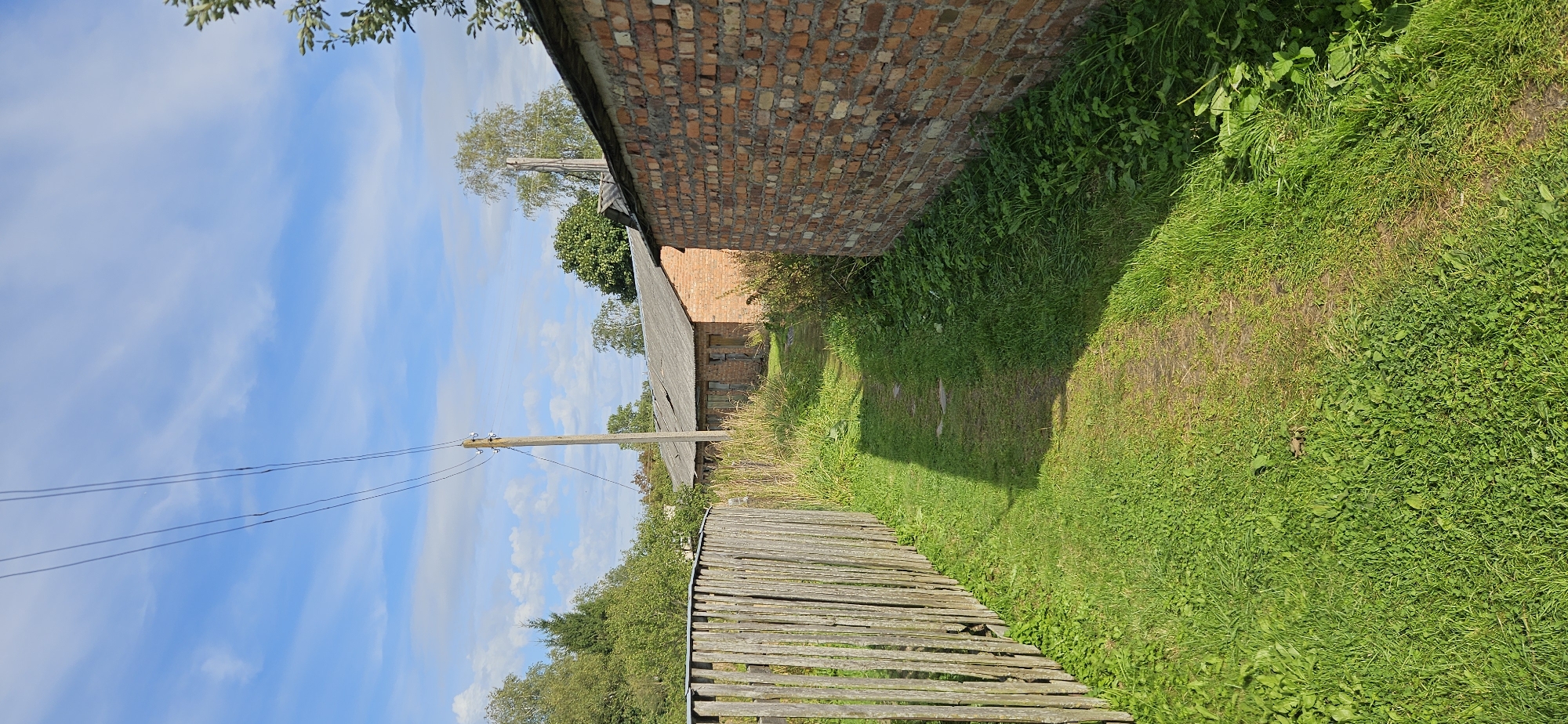 The image depicts an outdoor scene with a narrow dirt path bordered by a rustic wooden fence on the left and a weathered brick wall on the right. The path leads towards a long, low-lying building with a corrugated roof and multiple doorways. Overhead, telephone wires stretch across a bright blue sky with scattered white clouds. Green grass and various plants grow along the path and at the base of the walls and fence. Trees and foliage are visible in the background, surrounding the building and extending into the distance. The scene appears to be in an rural or semi-rural setting, possibly within the Мазалаўскі сельскі Савет, Belarus. There are no people or animals visible in the image. The lighting suggests it is daytime, with the sun casting shadows from the wooden fence onto the path.