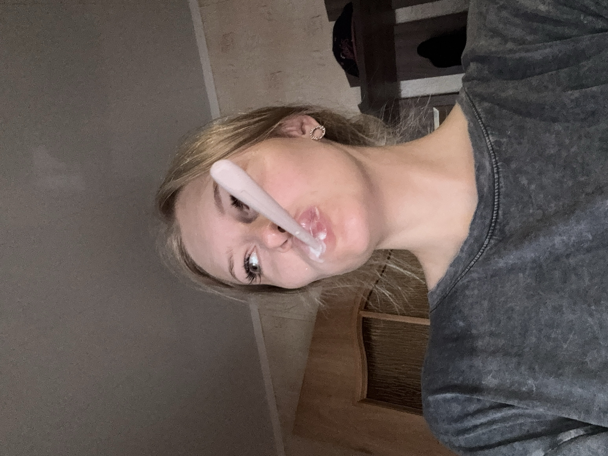 The image shows a close-up of a young woman's face as she is about to ingest something from a spoon. She has blonde hair and is wearing a grey t-shirt and a circular earring. Her mouth is open and her eyes are looking to the side. The background appears to be an indoor setting with a wooden door and furniture. There is no indication of the specific location within the city of Бородулино, Russia, or any crime scene details.