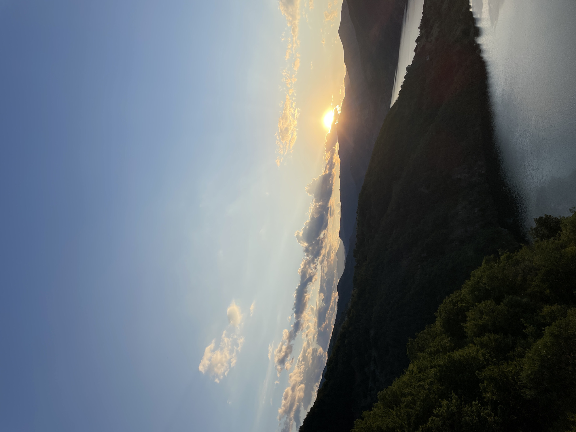 The image depicts a mountainous landscape at sunset. The sun is setting behind a range of silhouetted mountains, casting a warm glow on the scattered clouds. In the foreground, a dark, forested mountainside slopes down towards a body of water. The water, likely a lake or river, reflects the light of the sunset. The sky is a clear blue, transitioning to a softer hue near the horizon. The scene captures a serene natural environment, characteristic of the region around Borovitsa, Bulgaria. No people or specific man-made structures are visible.