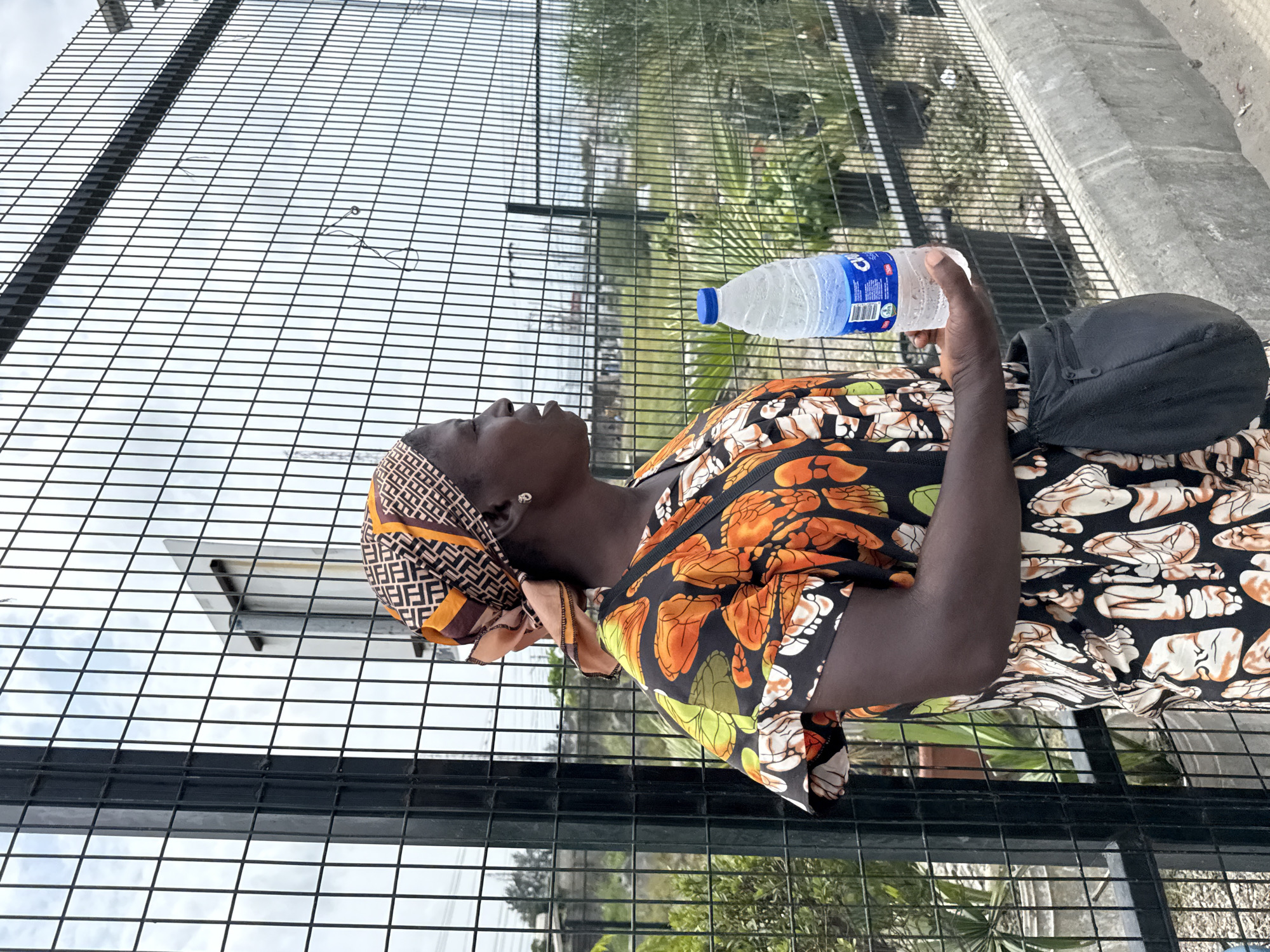 The image captures a person standing outdoors in Lekki, Nigeria, holding a plastic water bottle. The individual has dark skin and is wearing a colorful, short-sleeved top with patterns resembling orange and green leaves or abstract shapes on a dark background. They are also wearing a patterned headscarf in shades of brown, beige, and black, featuring a geometric design. A single silver stud earring is visible on their right ear. A black shoulder bag is slung over their right shoulder.
The person is holding a "Cway Pure Water" brand bottle with a blue cap in their right hand. They appear to be looking upwards or slightly to their left, with a neutral expression.
The setting features a tall, black metal grid fence in the foreground and midground, behind which lush green vegetation, including palm-like plants, can be seen. Through the gaps in the fence, a bright sky with some cloud cover is visible, suggesting daytime. To the right of the person, a grey concrete ledge or barrier is present. The overall scene suggests a public or semi-public outdoor area.