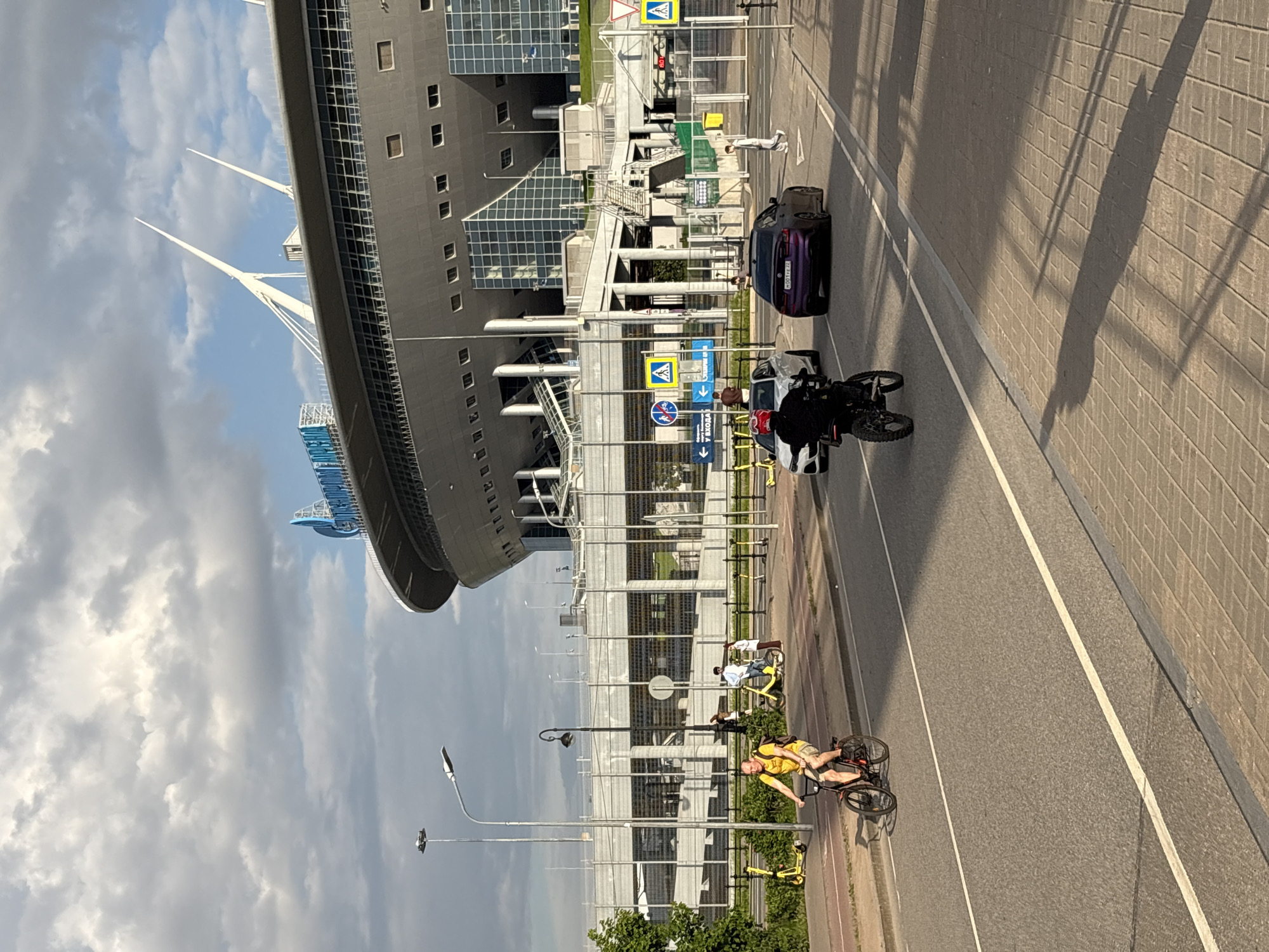 This image captures a vibrant urban scene in Saint Petersburg, Russia, on a partly cloudy day. The setting features a large, modern stadium-like building with a distinctive curved grey facade and multiple large, white, pointed mast structures reaching into the sky, suggesting a cable-stayed roof design. This building strongly resembles the Gazprom Arena (Krestovsky Stadium).
In the foreground and midground, a paved road with clear white markings runs parallel to the stadium, alongside a pedestrian or cycling path made of light-colored pavers. Sunlight casts long shadows from objects onto the road and a textured wall on the far right, indicating a specific time of day, possibly late afternoon or early morning.
Several subjects are visible on the road and path. A dark-colored car, possibly purple or very dark blue, is parked on the road, displaying a Russian license plate "H007EE 98". Behind it, a black and white motorcycle, possibly a police or security vehicle, is stopped, with a helmeted person sitting on it. Further back, a white car is partially visible.
On the adjacent path, a man in a yellow t-shirt and shorts rides a dark bicycle towards the viewer. Another person is seen on a yellow bicycle further down the path. Several yellow electric scooters and shared bicycles are parked along the side, suggesting recreational activity or local transport options. A person in a white tracksuit stands near the dark car.
Traffic signs are prominent, including a "pedestrian crossing" sign, a "road works ahead" sign, and blue directional signs, one indicating a mandatory turn right, and another with Russian text "У ВХОДА" (AT THE ENTRANCE). A traffic light shows the number "90", likely indicating seconds remaining. Streetlights and fencing line the area, with some green foliage. The scene depicts routine urban movement and leisure activities around a prominent architectural landmark.