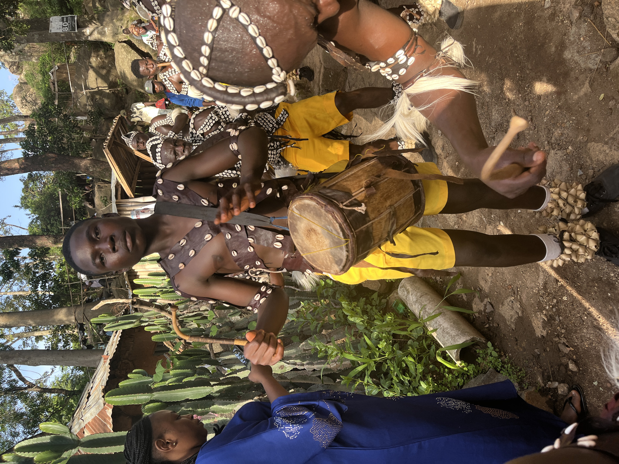 In Jos, Nigeria, a young man in traditional attire with shells and beads adorns his chest and arms, plays a drum. He wears yellow shorts and his ankles are decorated with shell ornaments. To his left, a woman in a blue dress, her hair braided, holds a wooden stick, possibly participating in the ritual. In the background, other individuals in similar traditional attire are visible, along with a rustic dwelling, large cactus plants, and a rocky, wooded landscape indicative of an outdoor gathering or ceremony.
