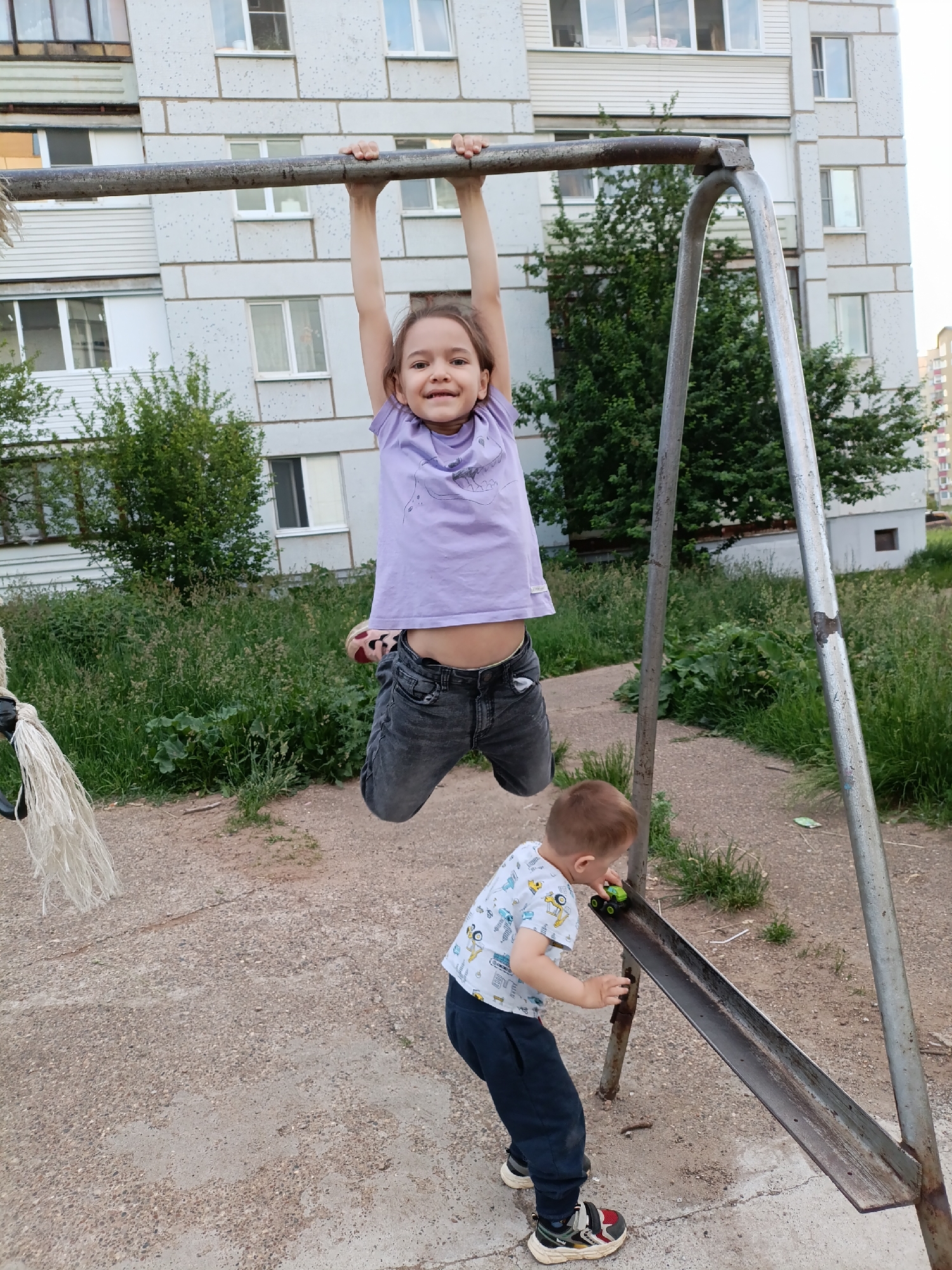 The image captures an outdoor scene in Chaikovsky, Russia, featuring two children on a playground in front of a multi-story residential building.
In the foreground, a pre-adolescent girl, approximately 5-8 years old, is suspended in mid-air, hanging from a horizontal, rusted metal bar with both hands. She wears a light purple short-sleeved t-shirt with a faint graphic, dark grey denim shorts, and light-colored shoes with red accents visible on her left foot. Her brown hair is parted and frames her face as she smiles directly at the camera. To the left of the bar, a bundle of white, frayed rope or fabric hangs downwards.
Below the girl and to her right, a younger boy, approximately 2-4 years old, stands with a slight forward lean. He is playing with a small, green toy car on an angled, rusted metal slide or ramp structure. He wears a white short-sleeved t-shirt with colorful vehicle patterns, dark blue track pants, and multi-colored sneakers.
The ground is a mix of uneven, light-colored concrete or compacted dirt with scattered pebbles, interspersed with patches of overgrown green grass and weeds. Behind the children and play structures, several medium-sized green leafy trees and bushes are visible. The background is dominated by a light-grey or white multi-story apartment building, at least five stories tall, characterized by numerous rectangular windows and some enclosed balconies. The building's facade shows visible seams, indicating pre-fabricated concrete panel construction. The sky above is light and overcast.
