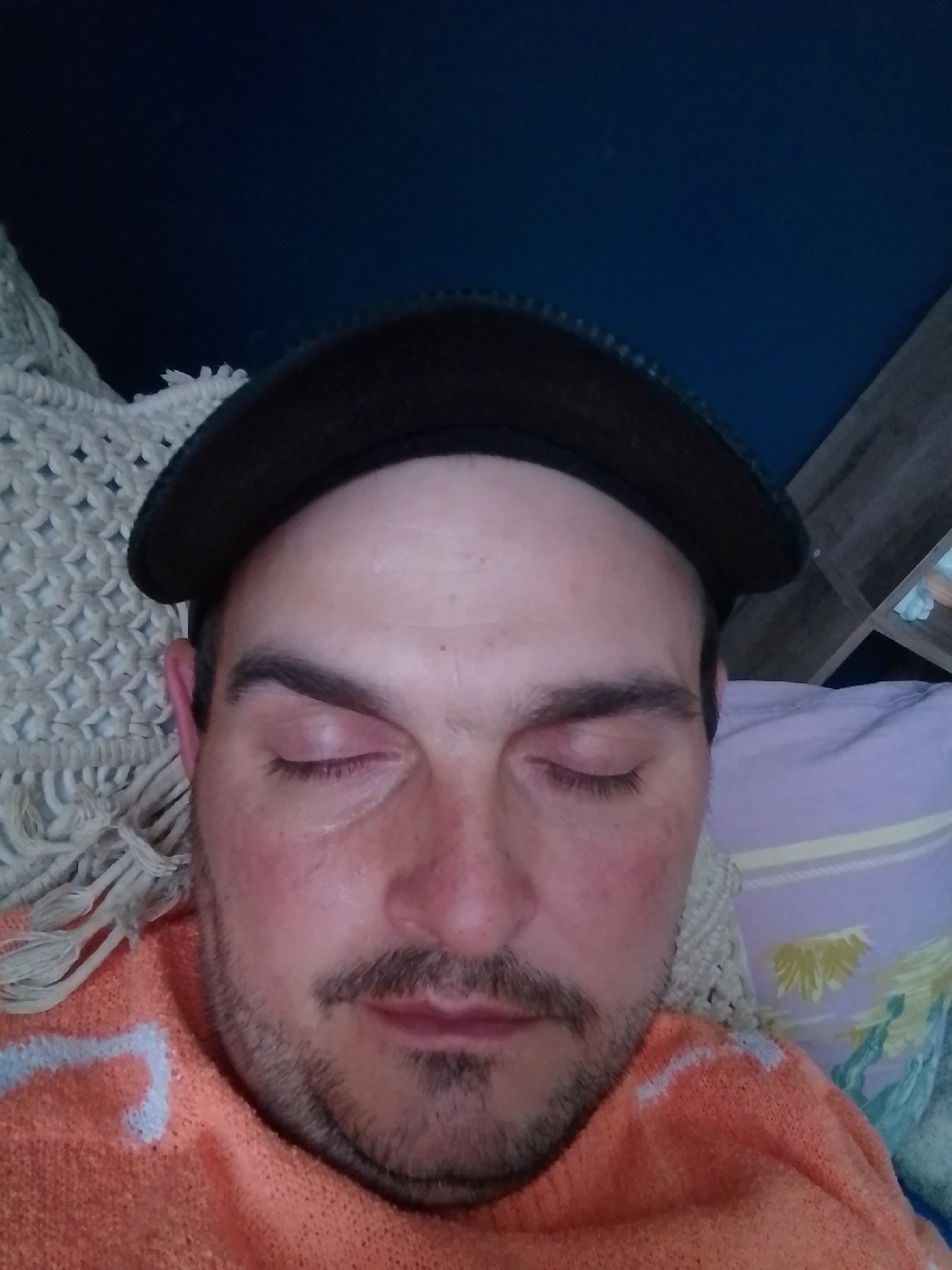 A close-up, overhead shot shows a person lying down, wearing an orange sweater with a blue and white pattern and a dark baseball cap. The person's eyes are closed, and they have a mustache and stubble. The background features a white crocheted blanket on the left and a purple pillow with a floral pattern on the right. To the right of the pillow, a wooden shelf unit is partially visible. The lighting is dim, with the primary light source appearing to be from the left. The image does not provide any specific information or context related to the city of Wedel, Germany.