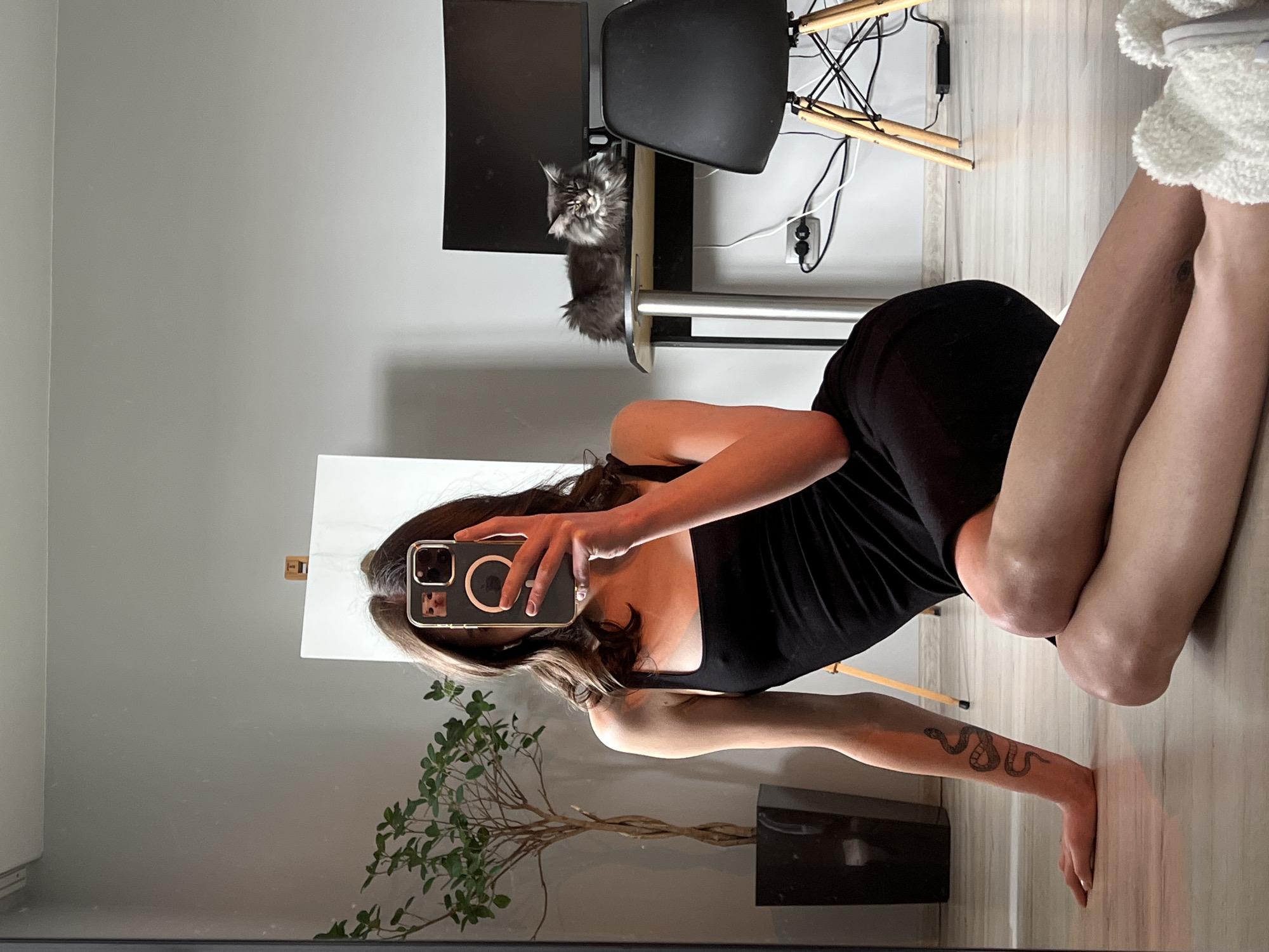 A young woman with brown hair is taking a mirror selfie indoors. She is wearing a black tank dress and sitting on a light-colored wooden floor. Her right arm has a snake tattoo. In the background, there is a plant in a black pot, an easel with a white canvas, a desk with a computer monitor and a cat, and a black chair with wooden legs. The cat is grey and fluffy, lying on the desk. There are power outlets and cords visible on the wall behind the desk. The room appears to be a residential living space, possibly in Ternopil, Ukraine, given the context.