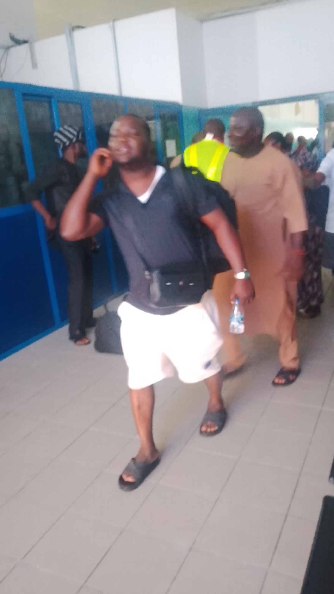 A medium shot shows a group of people indoors, possibly in a waiting area. The foreground is dominated by a man in a black t-shirt and white shorts, wearing black slides. He is holding a water bottle in his right hand and has a black satchel slung across his chest. He appears to be talking on his phone. To his right, a man in a tan-colored robe and black sandals is partially visible. In the background, other individuals are present, including a person wearing a yellow vest. Blue-tinted windows or panels line the left side of the scene. The floor is tiled with light-colored square tiles. The overall lighting suggests an indoor environment with some artificial illumination. The context of Orerokpe, Nigeria, is not discernible from the visual information alone.