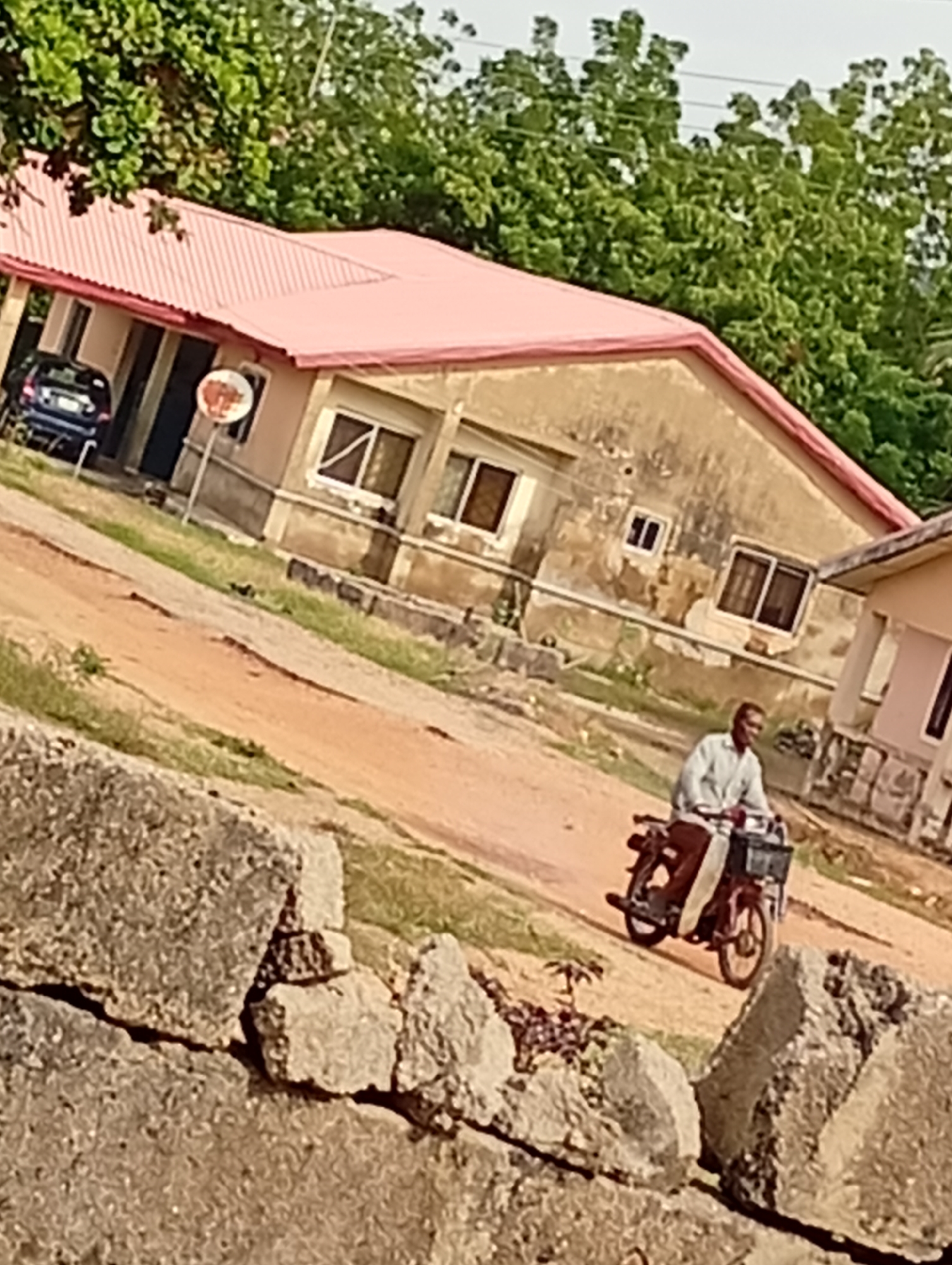 In an outdoor, daytime scene, a man is riding a motorcycle on a dirt road. The man is wearing a light-colored shirt and dark pants. The motorcycle is red and has a basket on the front. In the background, there is a building with a pink roof and several windows. Trees and vegetation are visible behind the building. In the foreground, large rocks are present. The overall scene suggests a rural or semi-urban environment.