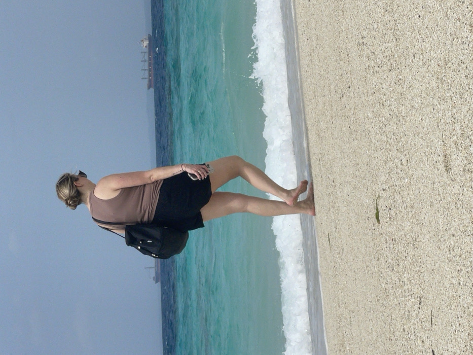 A female is depicted from a rear-right perspective, walking barefoot along a sandy beach in Zanzibar, Tanzania. She has light skin, blonde hair styled in a bun, and wears sunglasses, a sleeveless brown top, and black shorts. A black drawstring backpack is slung over her left shoulder, and she holds a light-colored smartphone in her right hand. Her left foot is lifted as she walks parallel to the waterline, where small white waves break against the light-colored sand. A small piece of green seaweed is visible on the sand in the foreground. The turquoise ocean extends into a deeper blue horizon under a hazy blue sky. Two large cargo ships are visible in the distant background; one is partially obscured on the left, and another, clearer vessel with multiple cargo holds is positioned further right.