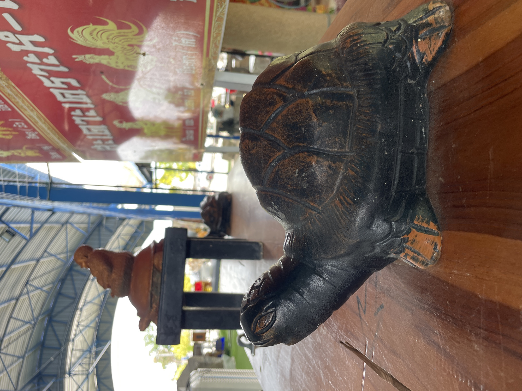 The image shows a detailed, dark-colored turtle figurine placed on a wooden surface. The figurine has a textured shell with an intricate pattern and painted details on its feet and head, including visible eyes. In the background, slightly out of focus, are other objects and structures. To the left, a sculpture of a seated figure on a turtle is visible on a dark stand. Further back, a red banner with yellow text and depictions of roosters is present, suggesting a location in Tambon Na Chom Thian, Thailand, possibly a market or a place of cultural significance. The environment appears to be an outdoor or semi-outdoor covered area, with a metal roof structure visible above.