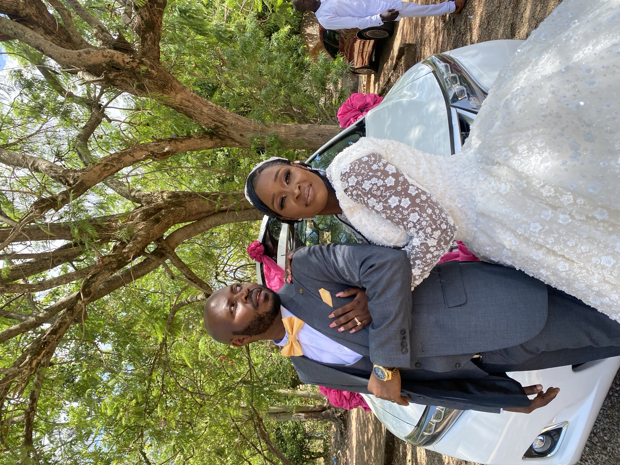 The image captures a wedding couple posing in front of a white car adorned with pink decorations. The groom, wearing a grey suit with a gold bow tie and a gold watch, stands next to the car, while the bride, in a white sequined wedding gown and headband, leans on him. They are both smiling at the camera. A tree with dense foliage is overhead, casting shadows on the scene. In the background, another car is visible, along with a person in a white outfit. The location is Dutsen Kato, Nigeria, as specified.