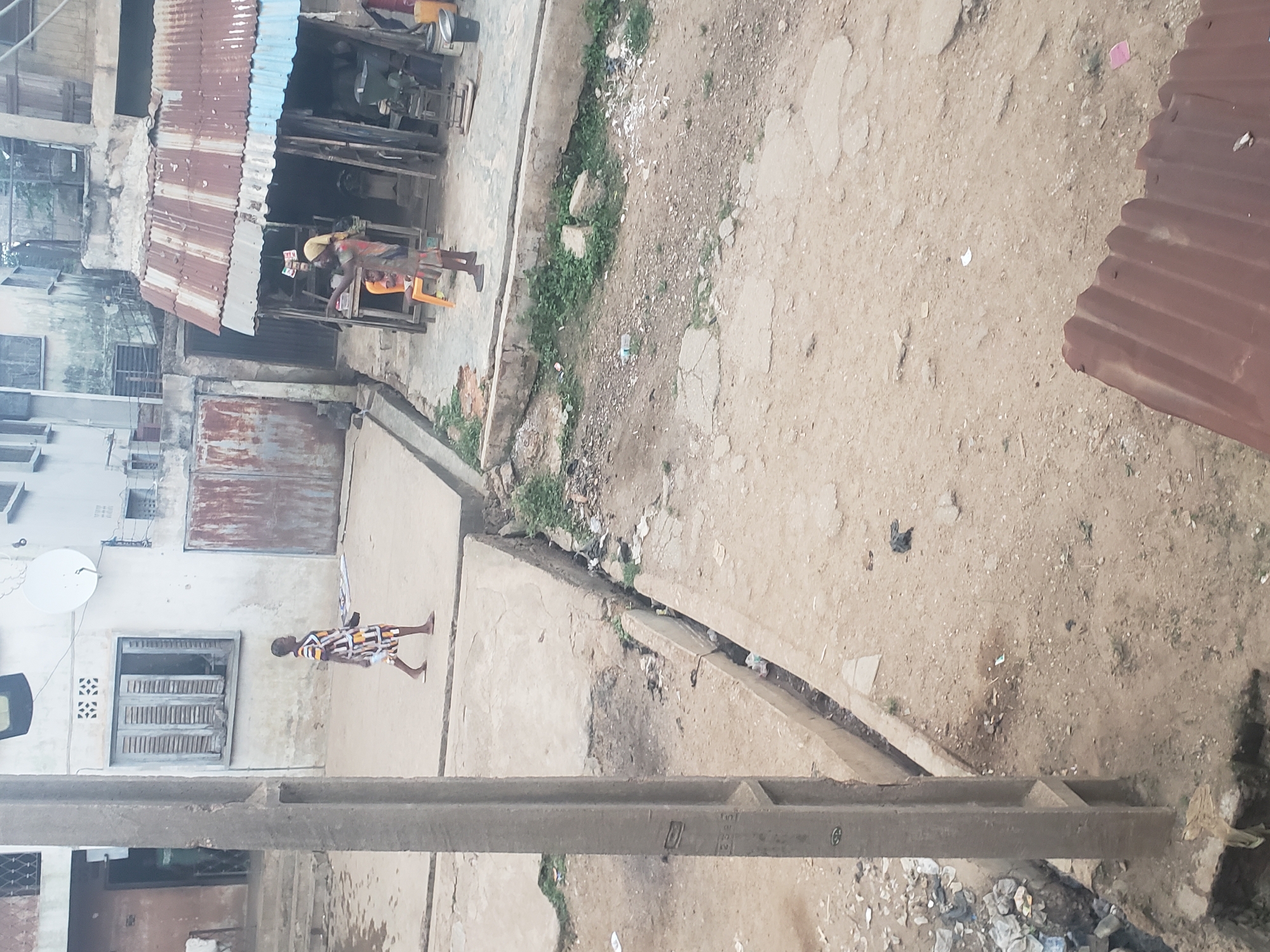 The image captures a street scene in Alalubosa, Nigeria. A dirt road is visible, with scattered debris and vegetation. On the left, there are buildings with corrugated metal roofs. One building appears to be a shop with a man standing inside. A person in a patterned dress is walking in the opposite direction on a paved area. A concrete structure runs along the bottom of the image, possibly a utility pole support.