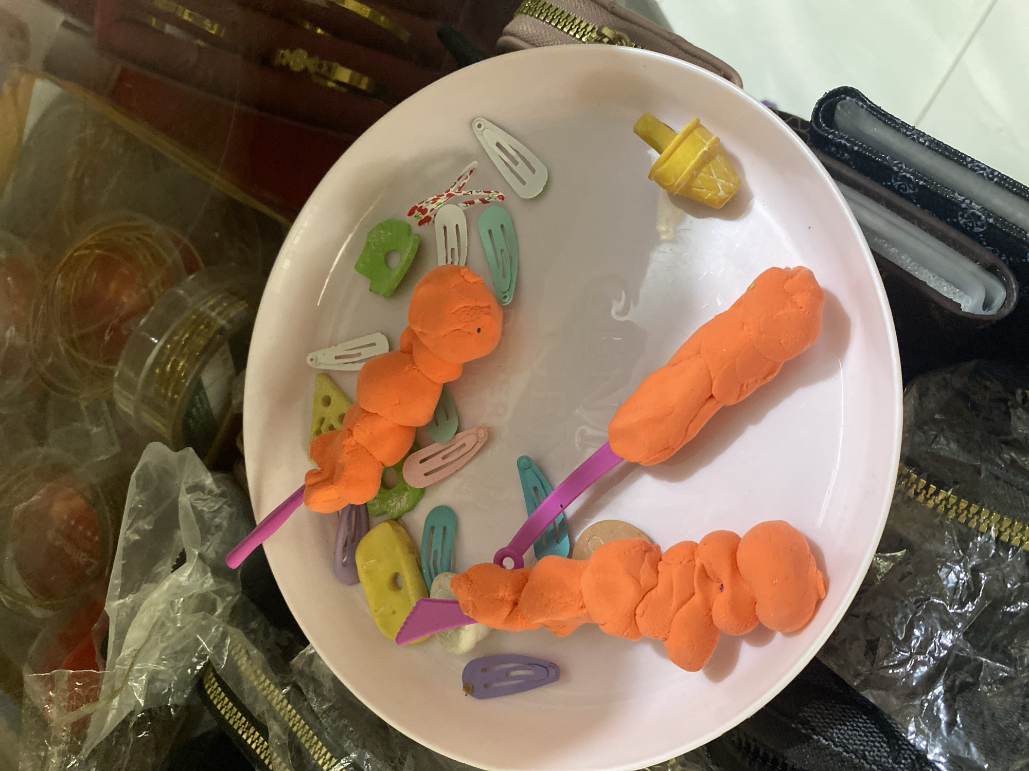 A close-up, overhead shot captures a pink plate filled with various objects. Two orange, Play-Doh-like creations are skewered on purple sticks, resembling food items. Scattered around them are colorful plastic hair clips in shades of purple, light blue, green, and pink, some with small red and white patterns. A miniature yellow ice cream cone toy rests near the bottom right of the plate. The plate sits on a dark surface, possibly a table or floor, with portions of black and metallic-gold textured materials visible in the background, suggesting the presence of bags or other accessories. The lighting is artificial, casting soft shadows and highlighting the textures of the objects on the plate. The scene appears to be an indoor setting, likely a domestic environment in Ta', Indonesia, given the context.