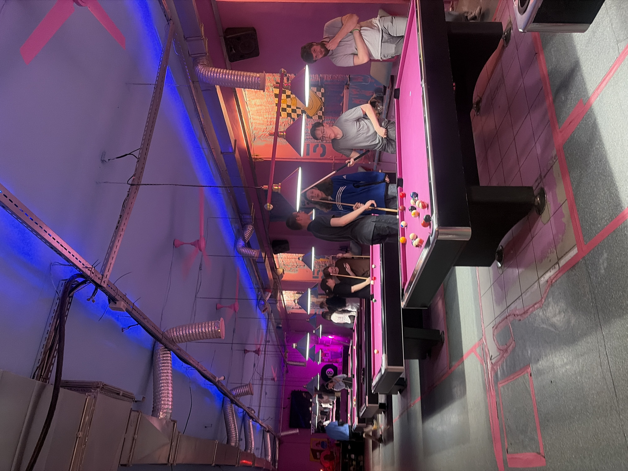 A group of young adults are playing billiards in a dimly lit establishment. The room is decorated with pink walls and purple lighting, creating a vibrant atmosphere. Several pool tables with pink felt are arranged throughout the space, with balls and cues visible on each. A few people are actively playing, while others observe or stand nearby. The ceiling features exposed ventilation ducts and pink ceiling fans, adding to the industrial-chic aesthetic. The floor is a dark gray with pink markings, possibly indicating designated playing areas. In the background, a large black "8" ball hangs on the wall. The scene suggests a casual and recreational environment, possibly a bar or entertainment venue.