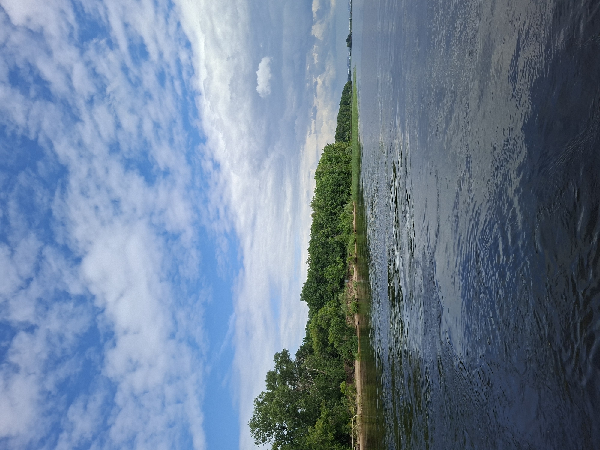 The image depicts a wide, calm body of water, likely a river or lake, with a lush green treeline along its bank in the distance. The sky is partly cloudy, with bright blue patches interspersed with fluffy white clouds and some darker, more ominous clouds towards the right side of the frame. The water in the foreground exhibits gentle ripples, reflecting the sky and the greenery. The treeline is dense and varied, with some taller trees and a sandy shore visible at the base. In the far distance, along the horizon, faint structures or a distant shoreline can be perceived. The overall impression is one of a serene natural landscape under a dynamic sky. This scene is consistent with locations along the Volga River in Kazan, Russia, such as the area near the Komsomolsky Park or along the embankment. There are no people or specific actions visible in the image.