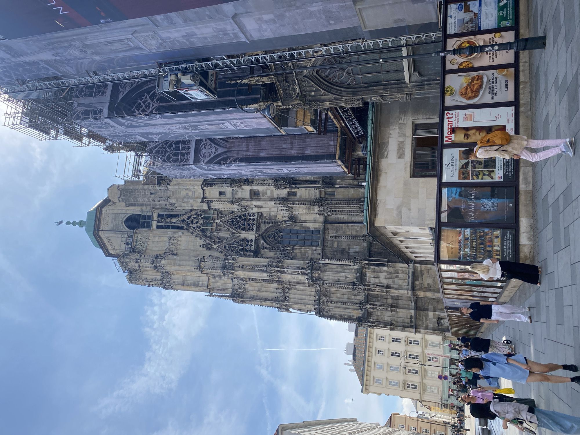 The image shows a wide shot of St. Stephen's Cathedral in Vienna, Austria, from a street-level perspective. The cathedral's iconic Gothic architecture is visible, with scaffolding and a large banner partially covering one of its towers, suggesting ongoing renovation work. In the foreground, a paved pedestrian walkway is lined with several large posters advertising various events or businesses. The posters display images of jewelry, shoes, a portrait of Mozart, and food. A number of people are walking on the sidewalk. One woman in the foreground is wearing a light-colored jacket and pink pants, carrying a tan backpack, and walking away from the camera. Other individuals are seen in the middle ground, some interacting, others walking alone. The sky is blue with scattered clouds and a faint contrail from an airplane. To the left of the cathedral, there are several buildings with traditional European architectural features, including windows and balconies. A lamppost stands on the right side of the frame, near the posters. The overall scene depicts a busy urban environment with a focus on historical architecture and public life.